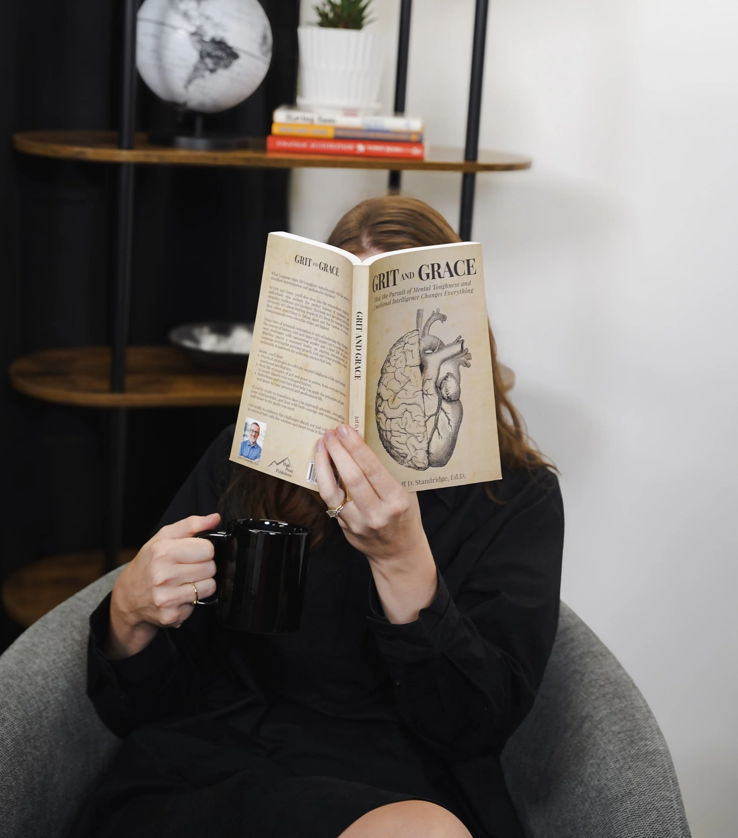 A person with long brown hair sitting on a gray couch, reading a book titled 'Grit and Grace' with an illustration of a heart and brain on the cover, holding a black mug.