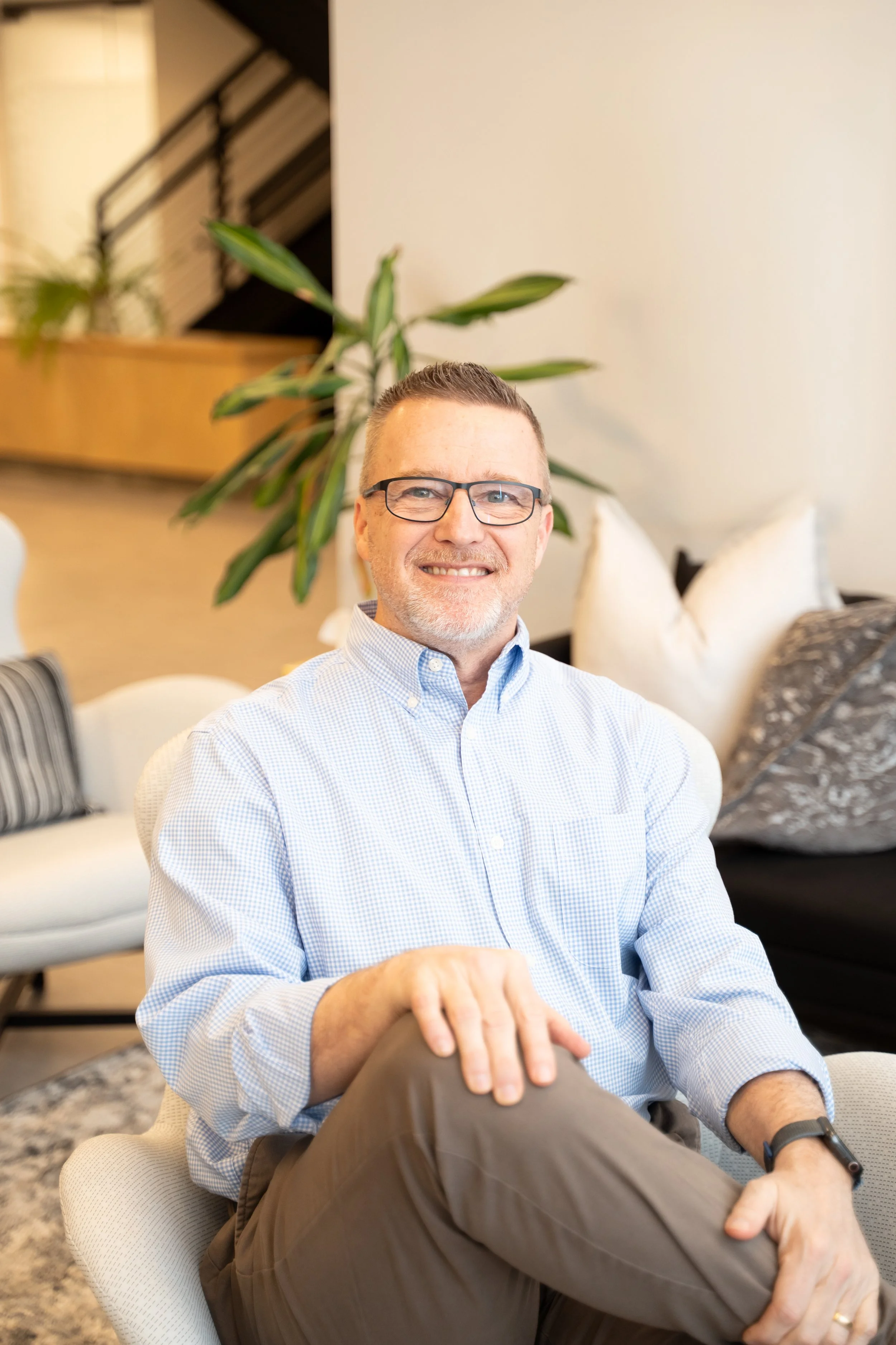 A man with glasses and a light blue dress shirt smiling while sitting on a white chair in a cozy room with pillows and a large plant in the background.