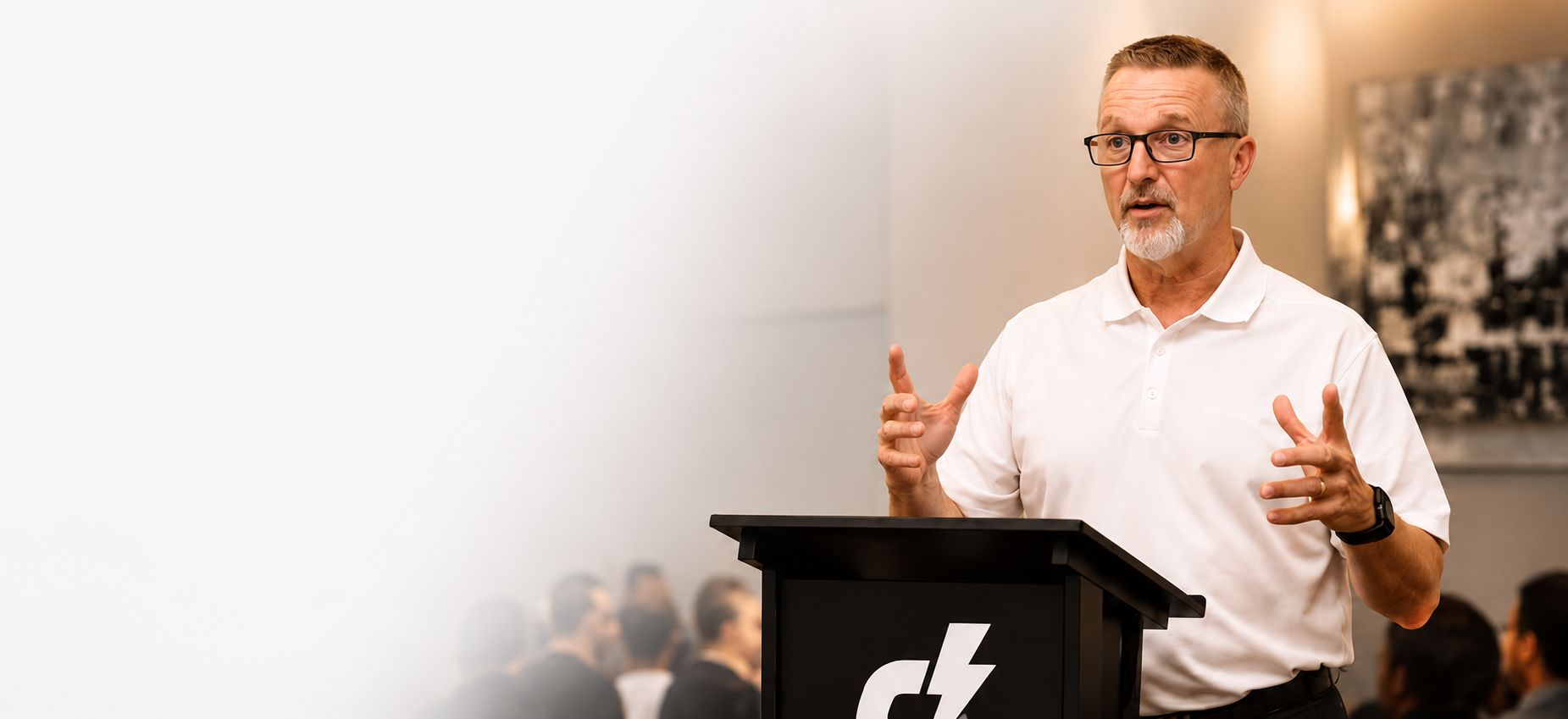A middle-aged man with glasses and a beard, wearing a white polo shirt, is speaking at a podium during a presentation or conference, surrounded by an audience.