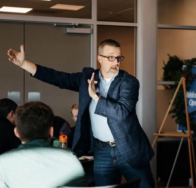 A man with glasses in a plaid blazer speaks passionately to a group of people in an indoor setting, using hand gestures.
