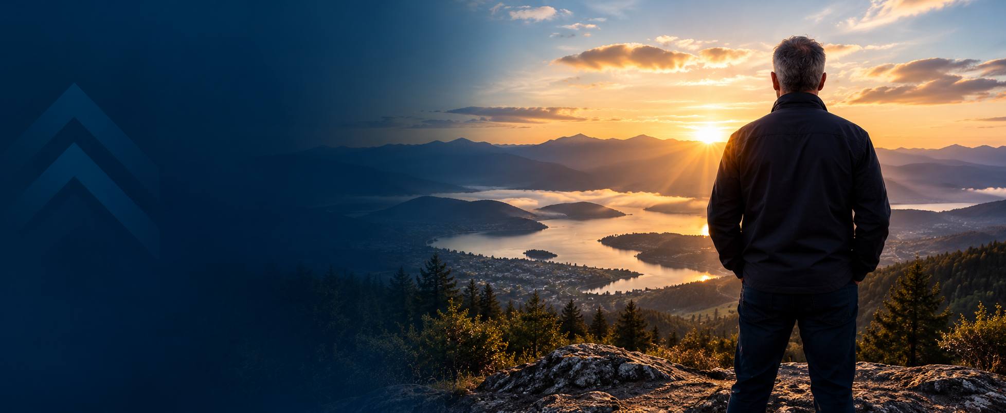 A man standing on a rocky ledge overlooks a lake and surrounding mountains at sunrise, with clouds overhead and sunlight breaking through.