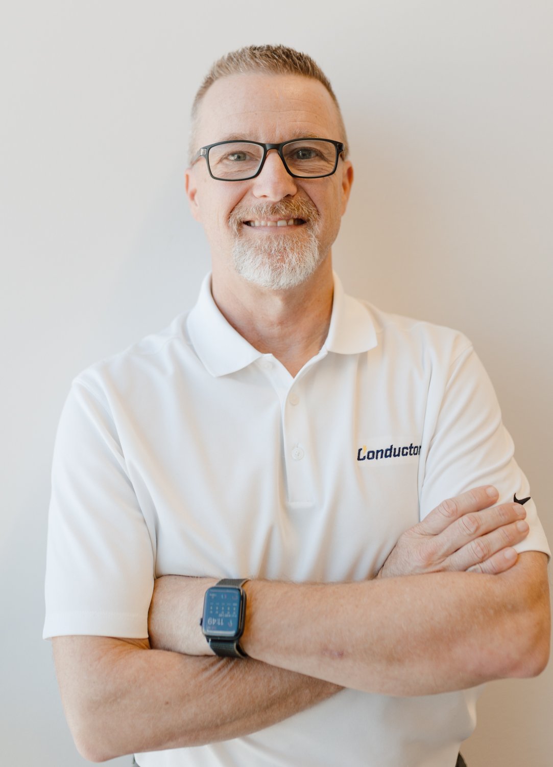 Portrait of a smiling man with glasses and a beard, wearing a white polo shirt with a logo that says 'Conductor,' standing with arms crossed against a plain background.