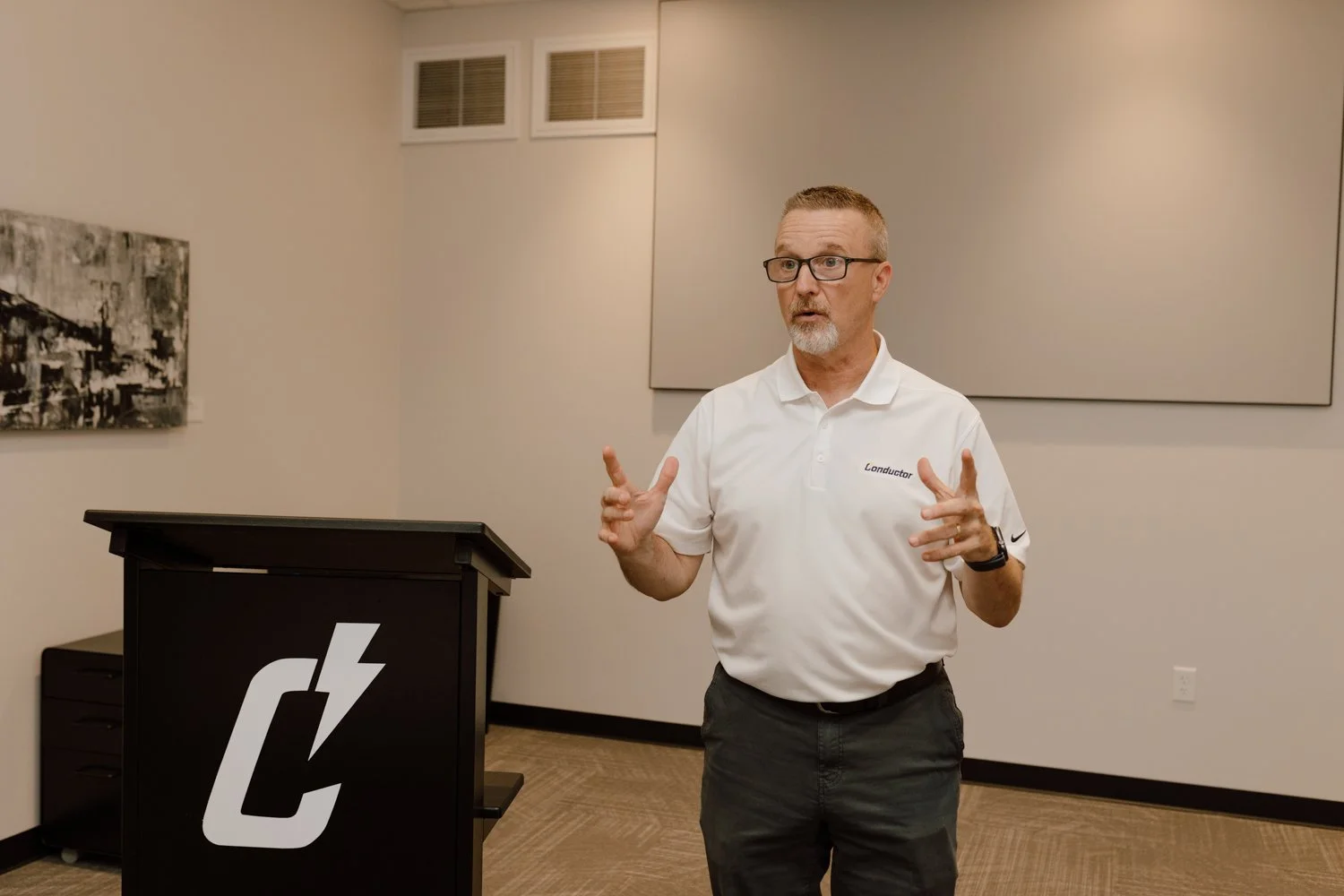 A man wearing glasses and a white polo shirt with the word 'L AND' is standing in a room, gesturing with his hands, and speaking. There is a black podium with a logo and a lightning bolt on it in front of him. The room has beige walls and two large acoustic panels.