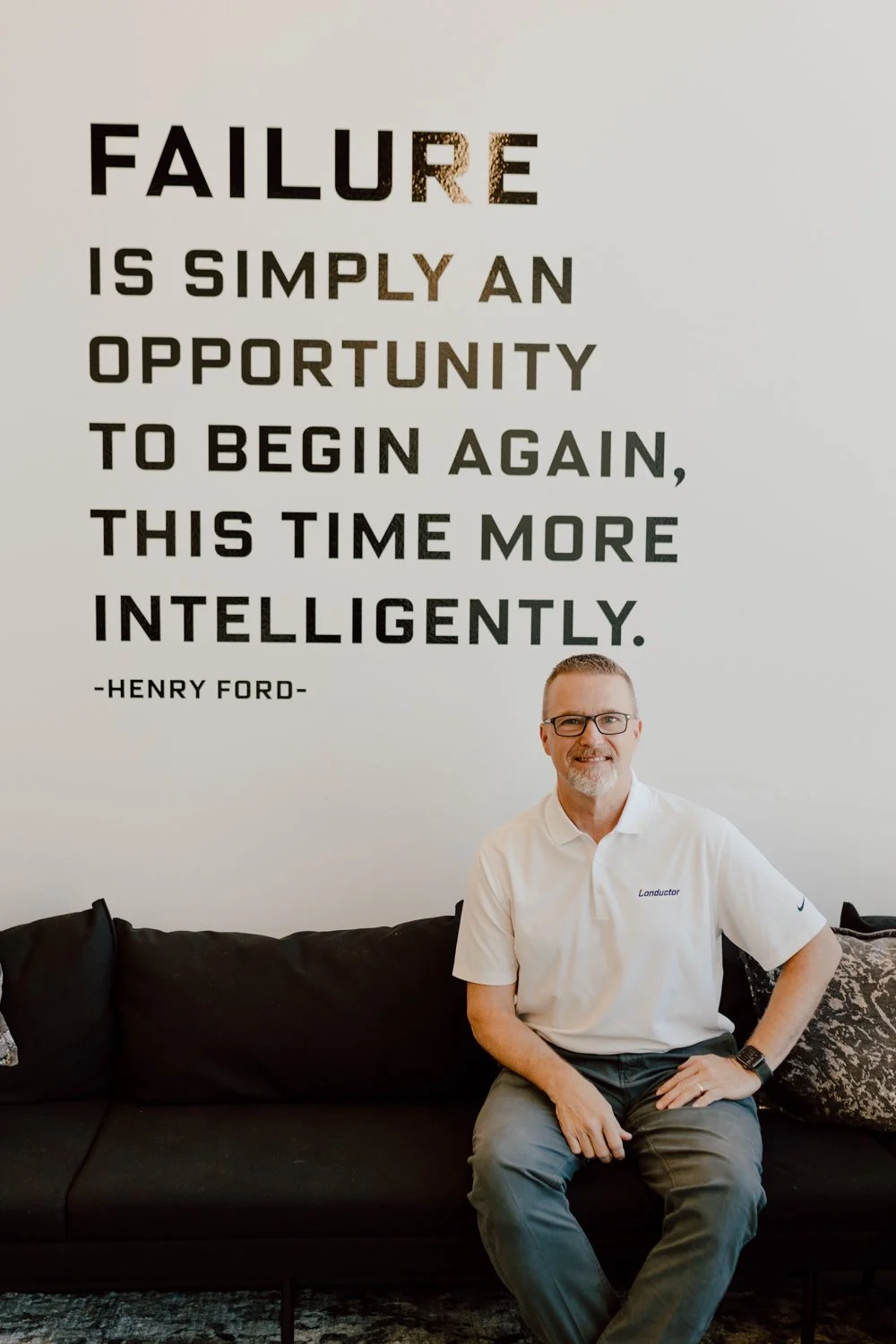 A man with glasses and a white polo shirt sitting on a black couch in front of a wall with a Henry Ford quote. The quote reads: 'Failure is simply an opportunity to begin again, this time more intelligently.'