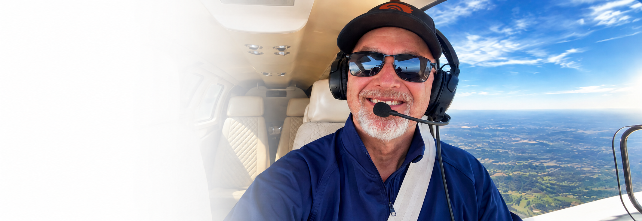 A man with a gray beard and sunglasses taking a selfie inside a small plane, wearing a headset with a microphone, blue jacket, and a cap. The background shows blue sky with some clouds and a landscape of fields below.