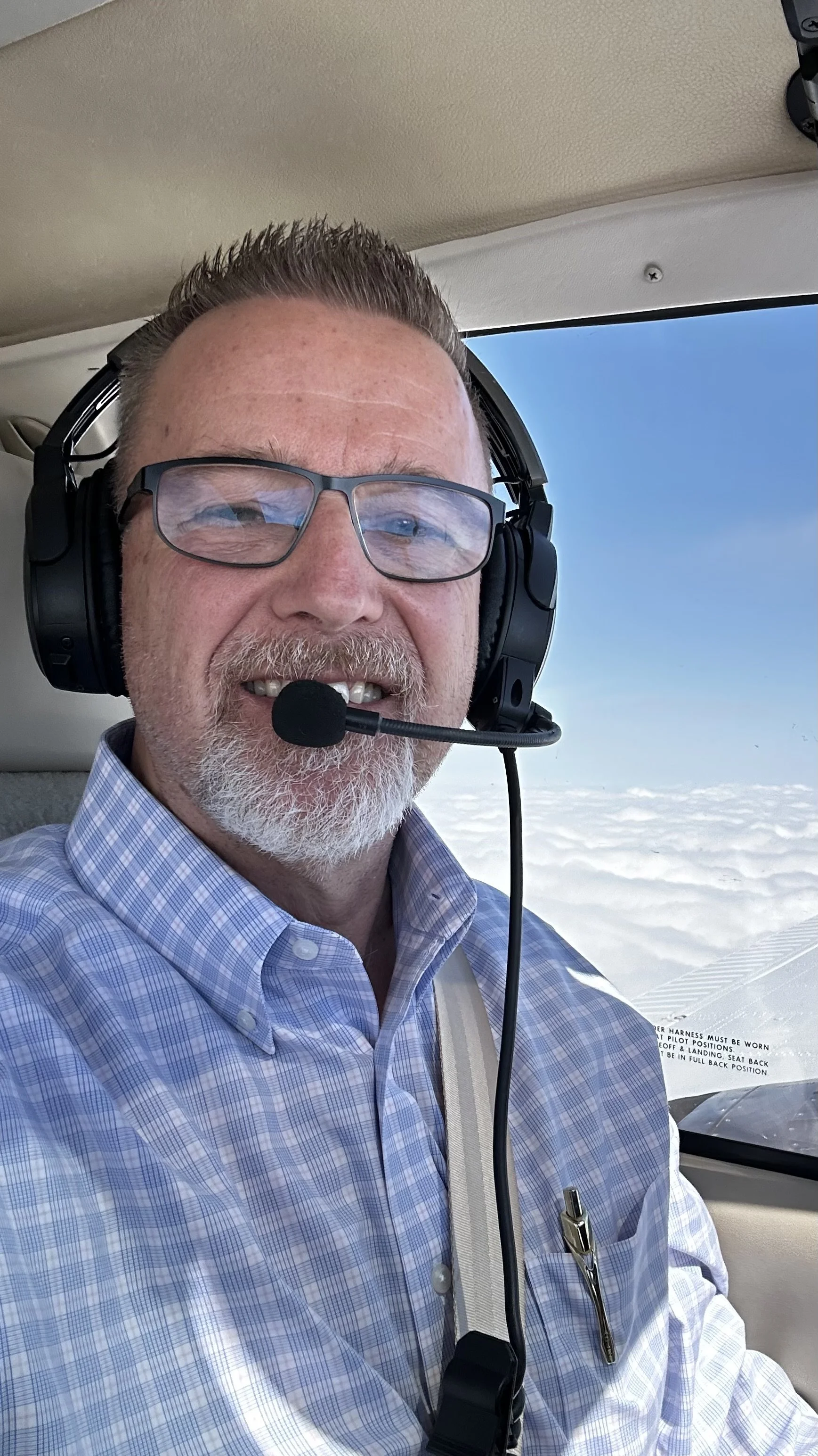 Man with glasses, beard, and mustache wearing a checkered shirt and headset, sitting in an airplane cockpit with blue sky and clouds outside.