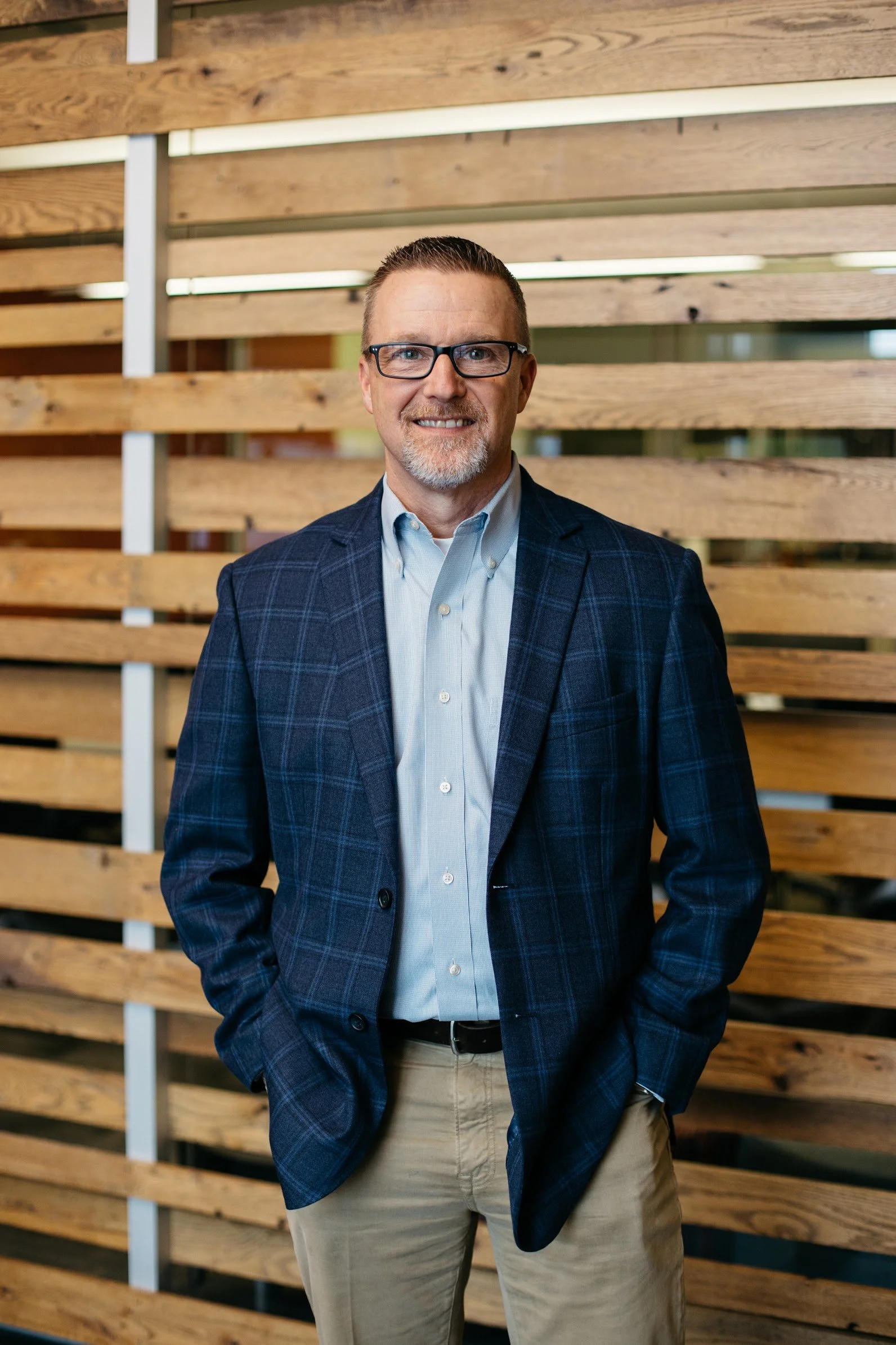 A middle-aged man with glasses, short hair, and a beard, smiling and wearing a blue plaid blazer over a light blue shirt, standing in front of a wooden wall with horizontal slats.