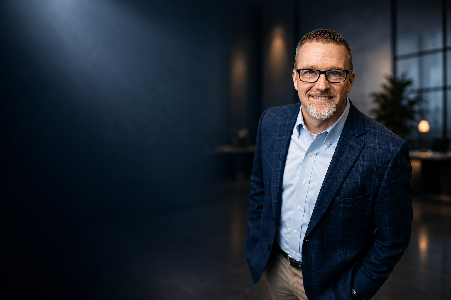 Smiling middle-aged man with glasses, wearing a blue plaid blazer and light blue shirt, standing in a modern office space with dark blue walls and large windows.