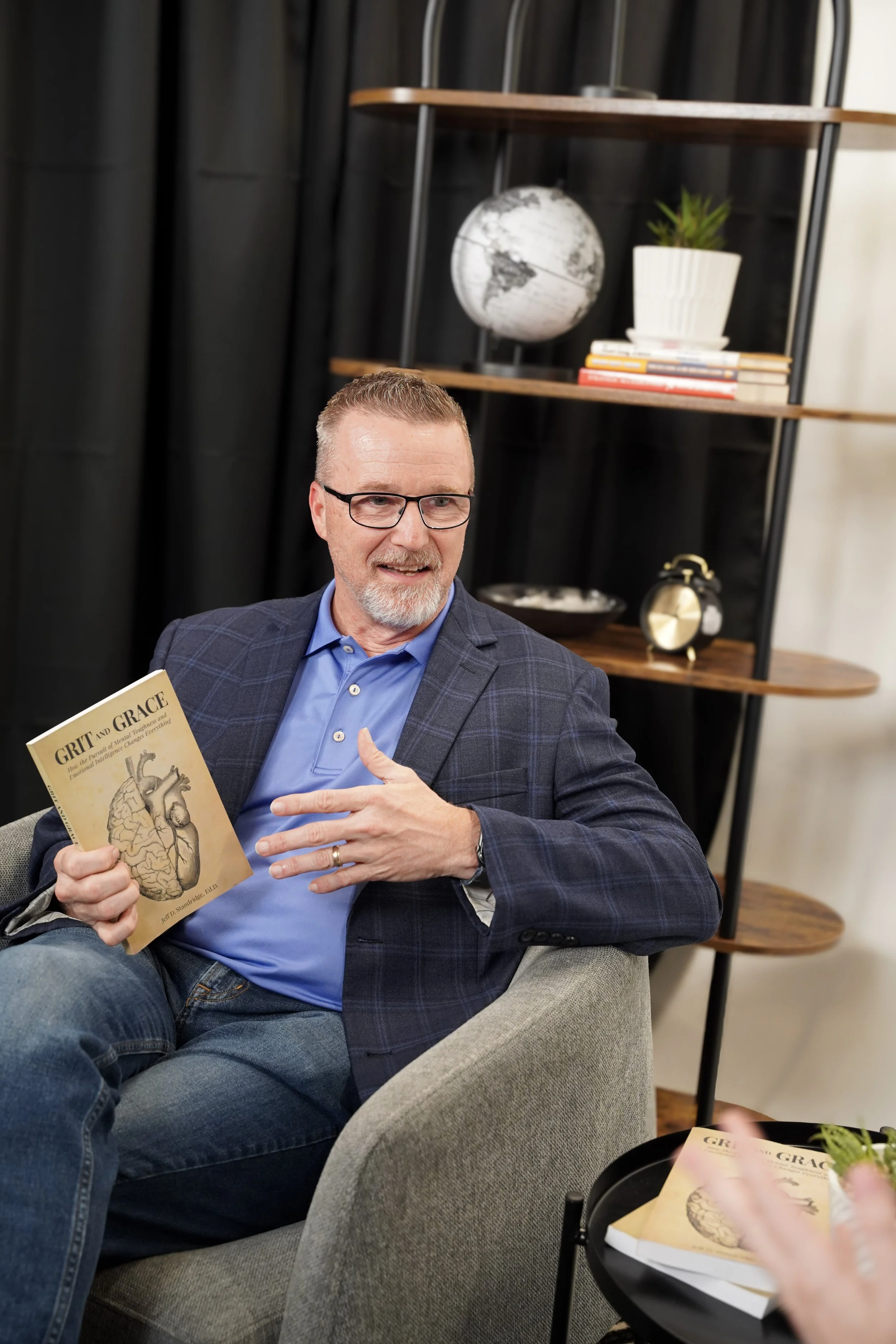 A man with glasses and a beard wearing a blue shirt and checkered blazer sitting on a light gray sofa, holding a book titled 'GRIT and GRACE' with a picture of a brain, in a room with black curtains and a modern shelving unit behind him.