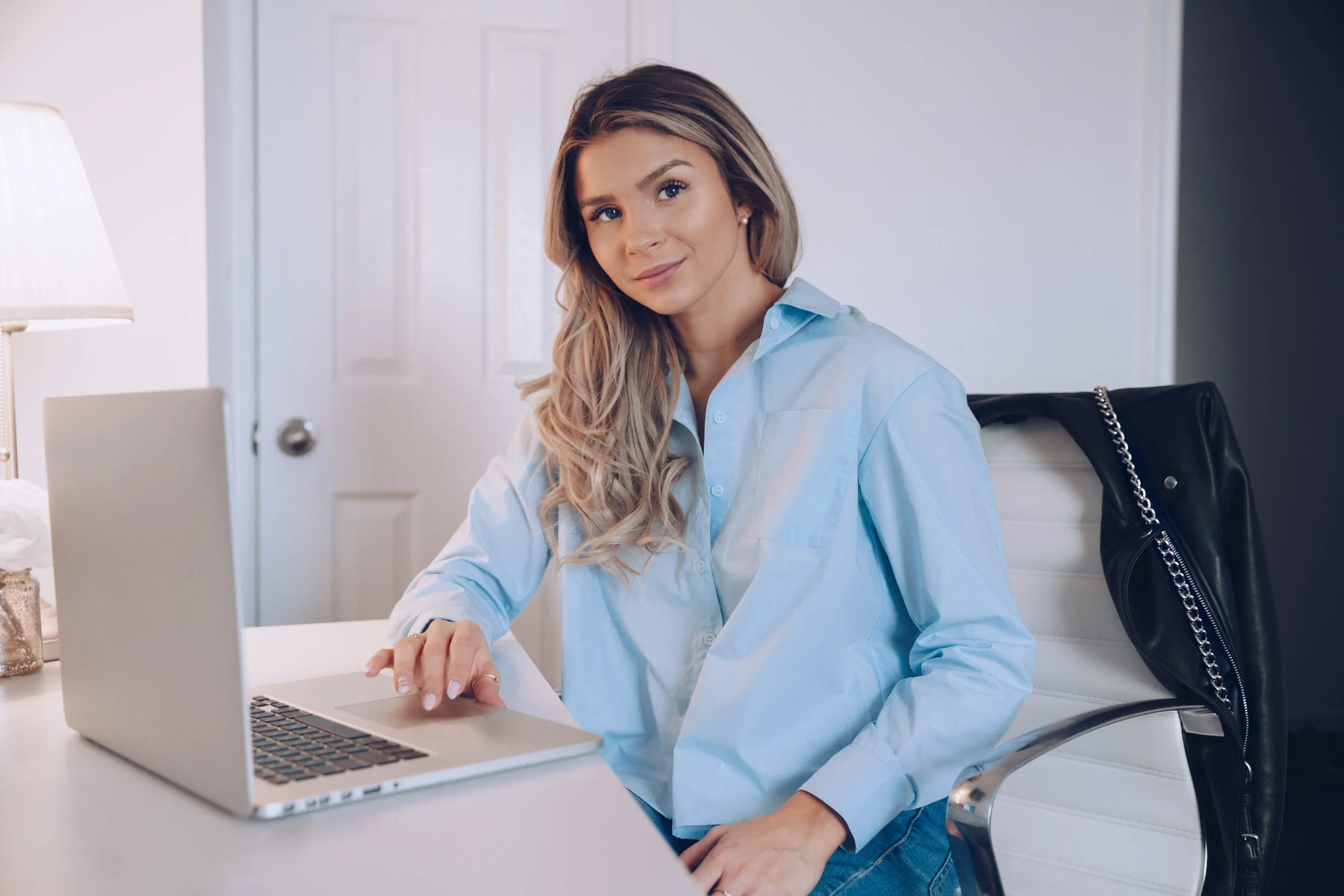 Young woman sitting at a desk with a silver laptop, looking at the camera, in a bright room with a white door, wearing a light blue button-up shirt.
