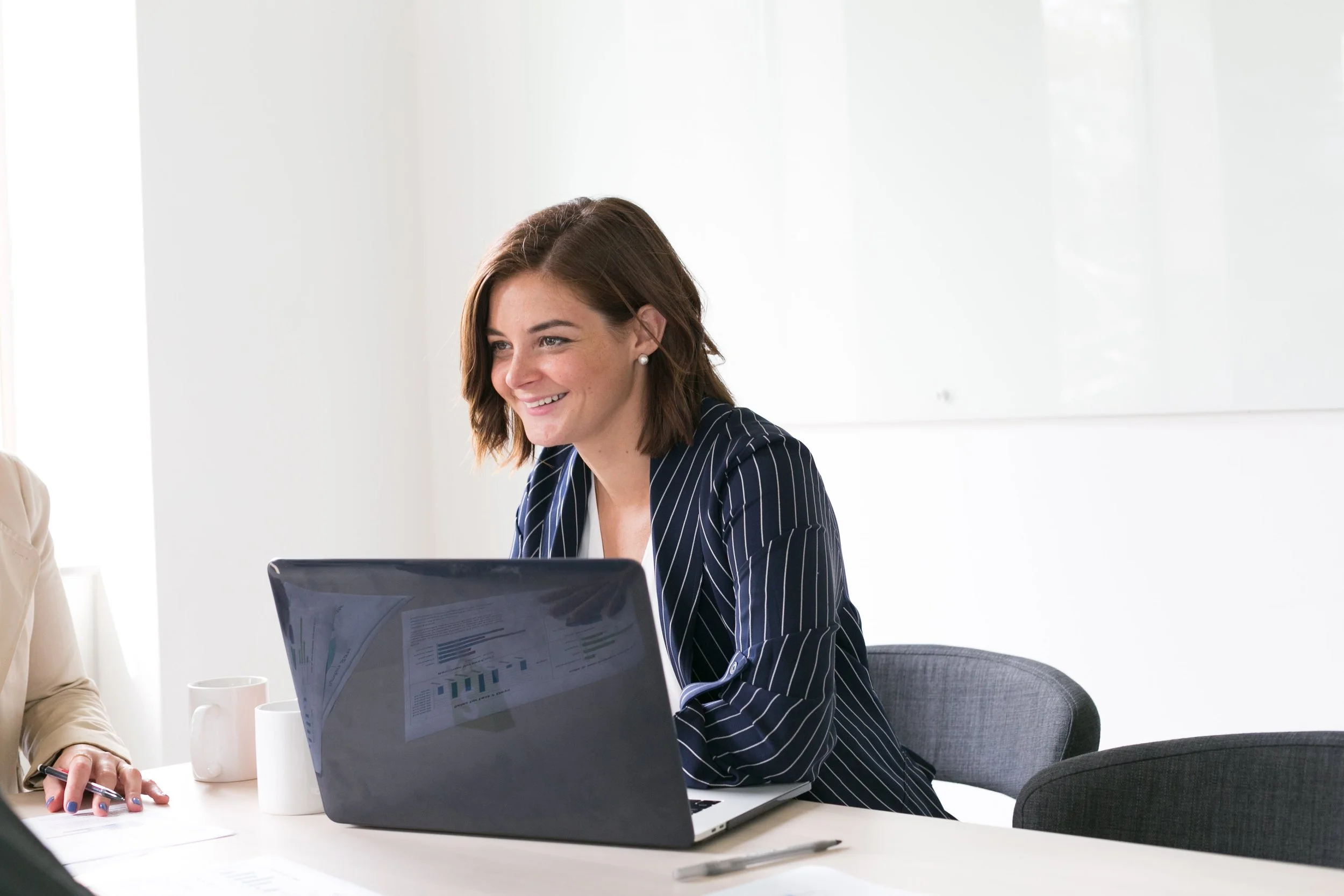 A woman with short brown hair, wearing a dark striped blazer, sitting at a desk with a laptop, smiling during a meeting.