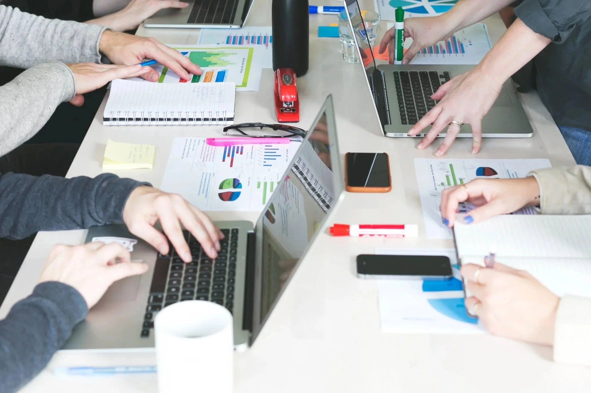 A group of people working at a shared table with laptops, documents, and office supplies, including pens, notepads, and charts.