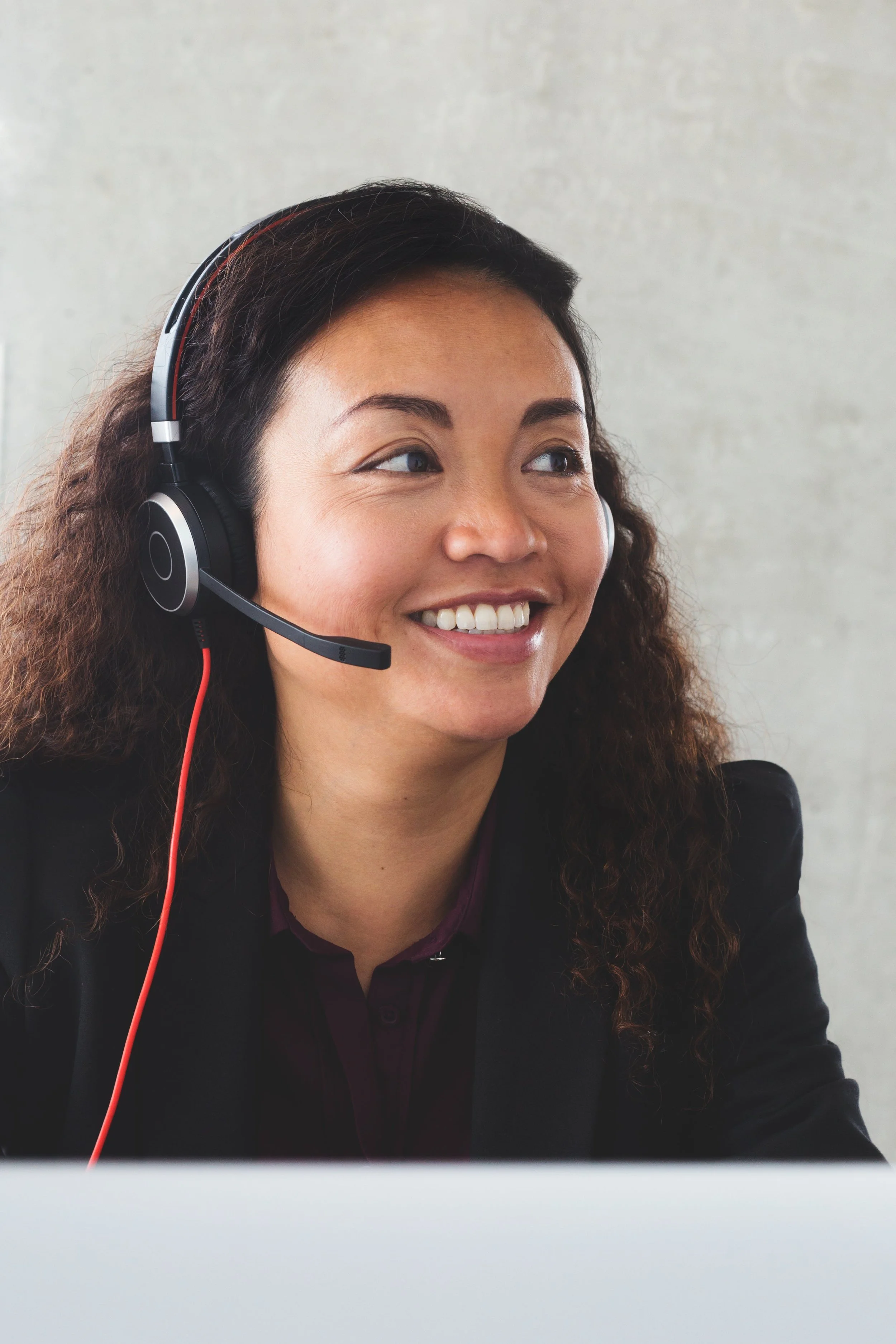 A woman with curly hair smiling and wearing a black headset with a microphone, sitting in front of a computer.
