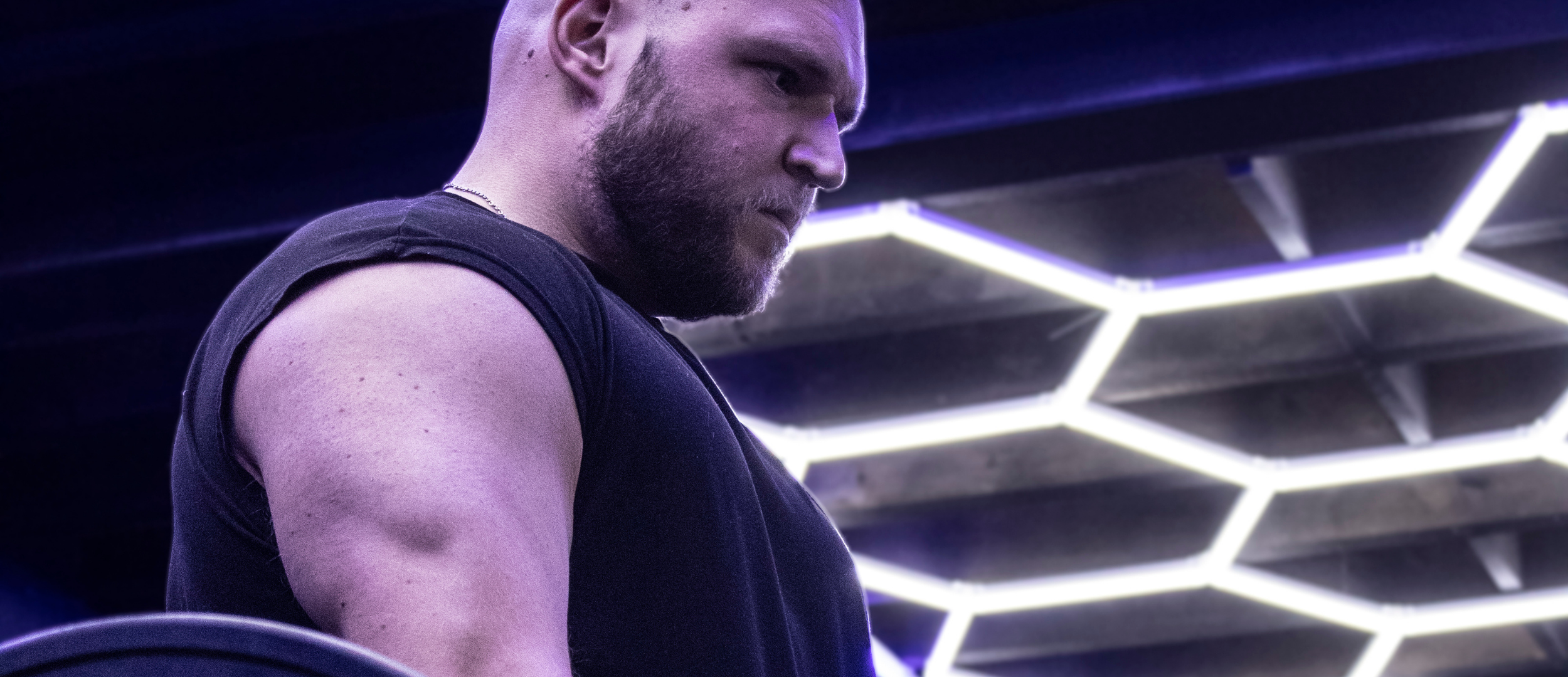 A man with a beard and muscular arm in a black sleeveless shirt, looking down, in a room with hexagonal LED lights on the ceiling.