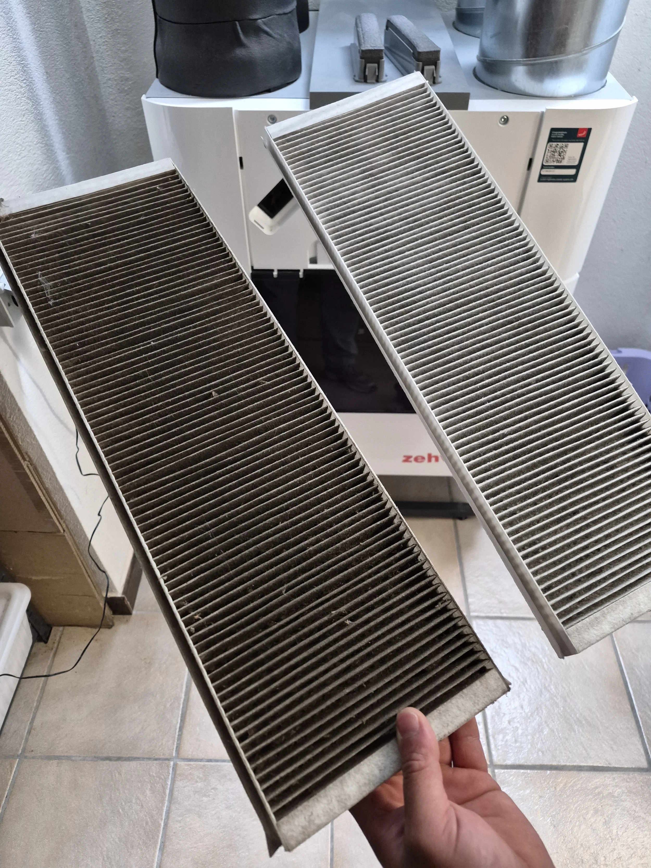 Person holding a dirty air filter in front of a cabinet with additional filters and cleaning tools.