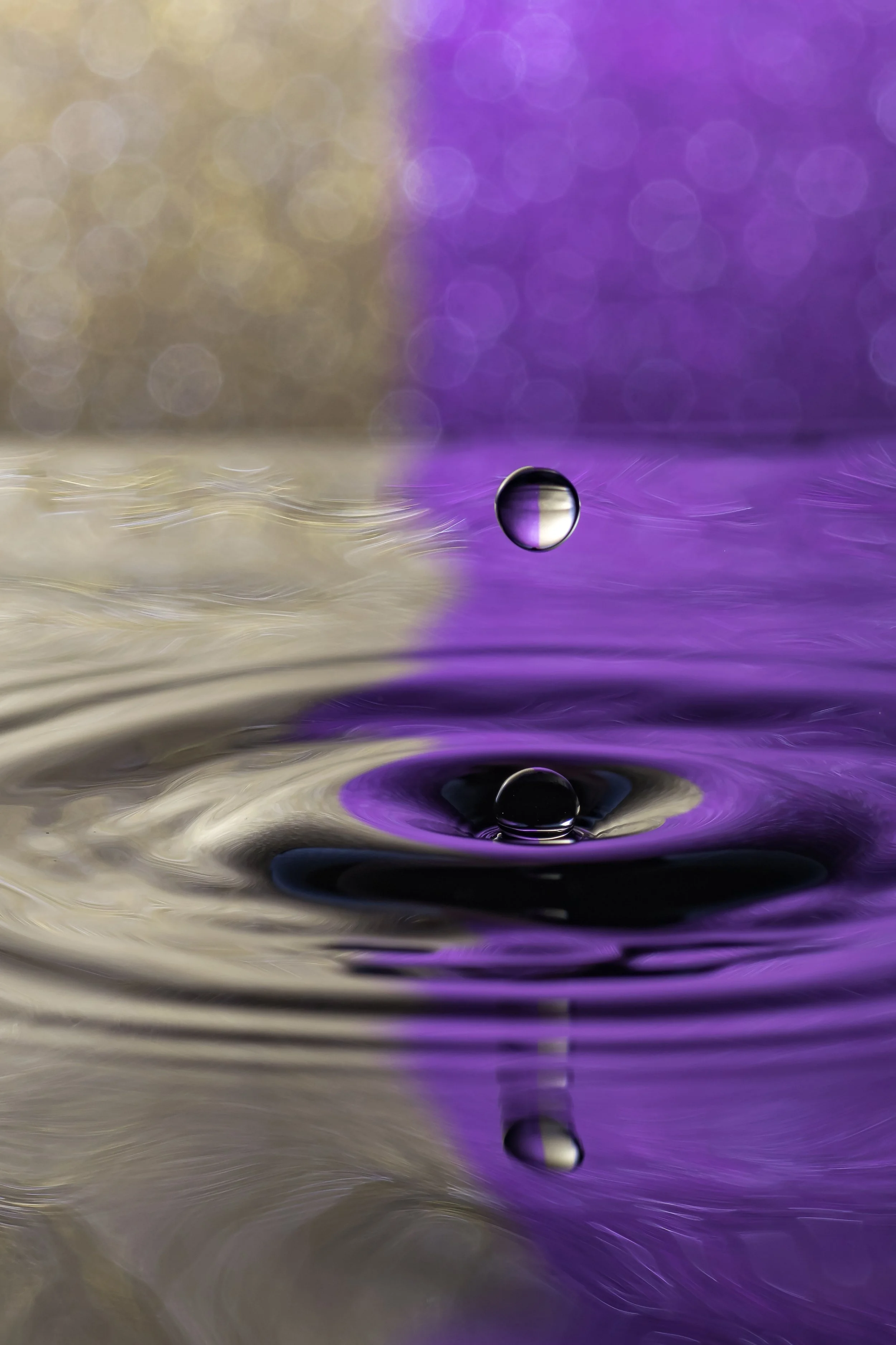 Close-up of a water droplet falling into a liquid surface, creating ripples with a purple and beige bokeh background.