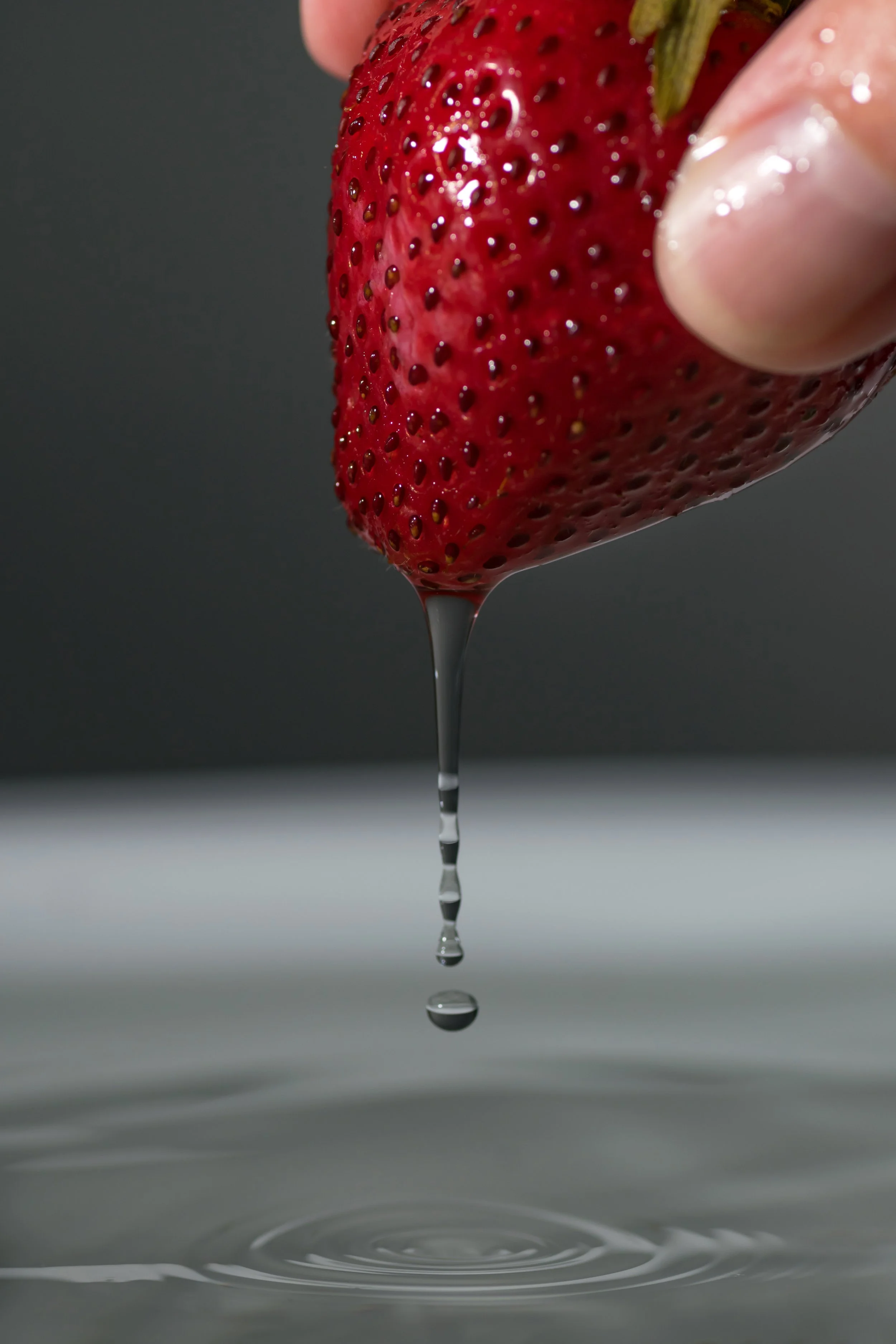 Close-up of a fresh strawberry being held, with honey or syrup dripping from it into a puddle below, against a dark background.