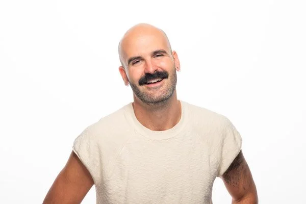 A brightly-lit headshot of Dylan smiling against a white backdrop. He is wearing a beige sweater, and has a mustache with a shaved head.
