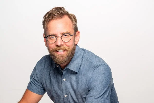 A brightly-lit headshot of Forty smiling against a white backdrop. He is wearing a blue short-sleeved button-up, round blue glasses, and has a bristled beard and coiffed blonde hair parted to one side.