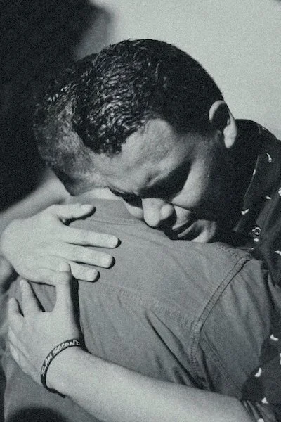 Black and white close-up of two men embracing tightly, one resting their face on the other's shoulder.