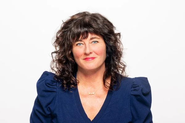 A brightly-lit headshot of Love smiling against a white backdrop. She is wearing a navy blouse and gold necklace, and has dark brown, shoulder-length curly hair.
