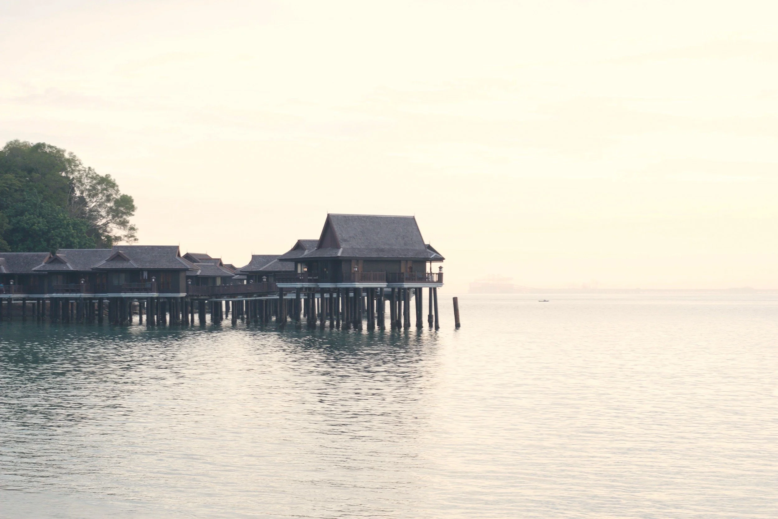 Traditional Malaysian wooden bungalows on stilts above calm tropical waters, surrounded by lush greenery.
