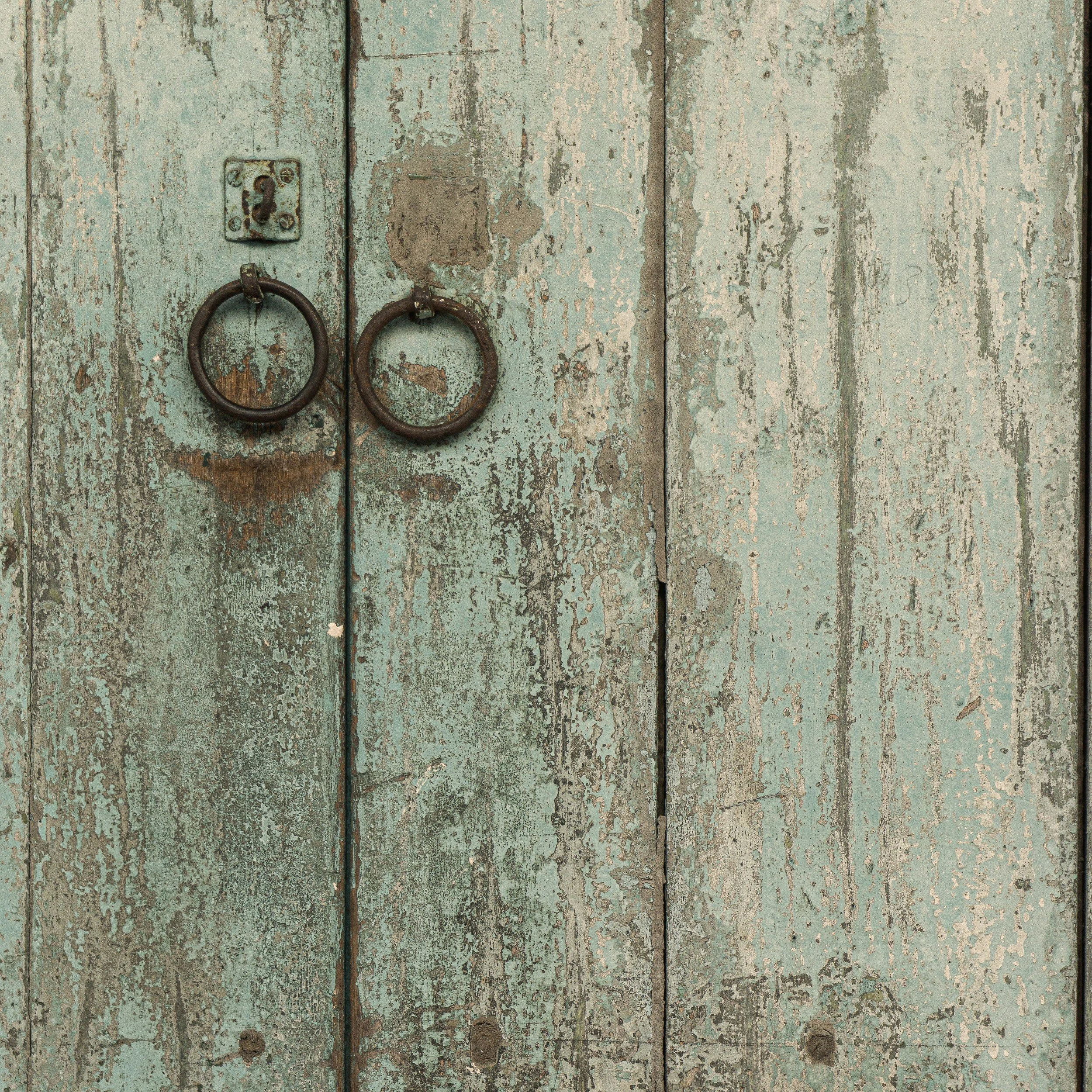 Close-up of an old Malaysian weathered wooden door with peeling mint green paint, rusted metal hooks, and a keyhole plate.