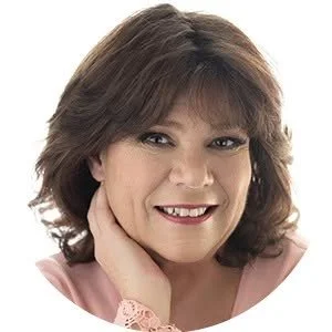 A woman with medium-length brown hair smiling and holding her chin with her hand, wearing a pink top.