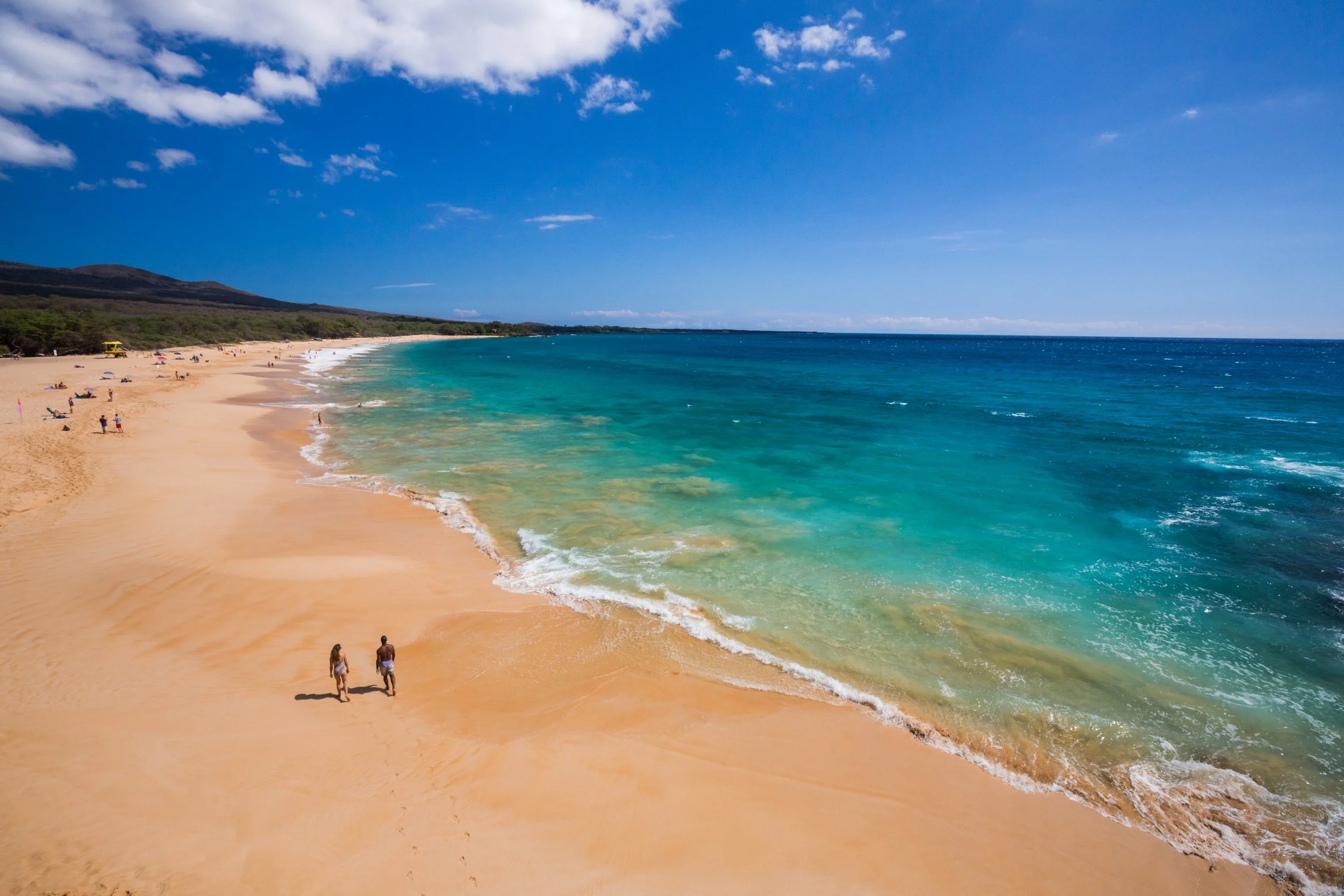 A scenic beach with golden sand, clear turquoise water, and a bright blue sky with scattered white clouds. People are walking along the shoreline and relaxing on the beach, with a distant mountain or hill covered in greenery in the background.