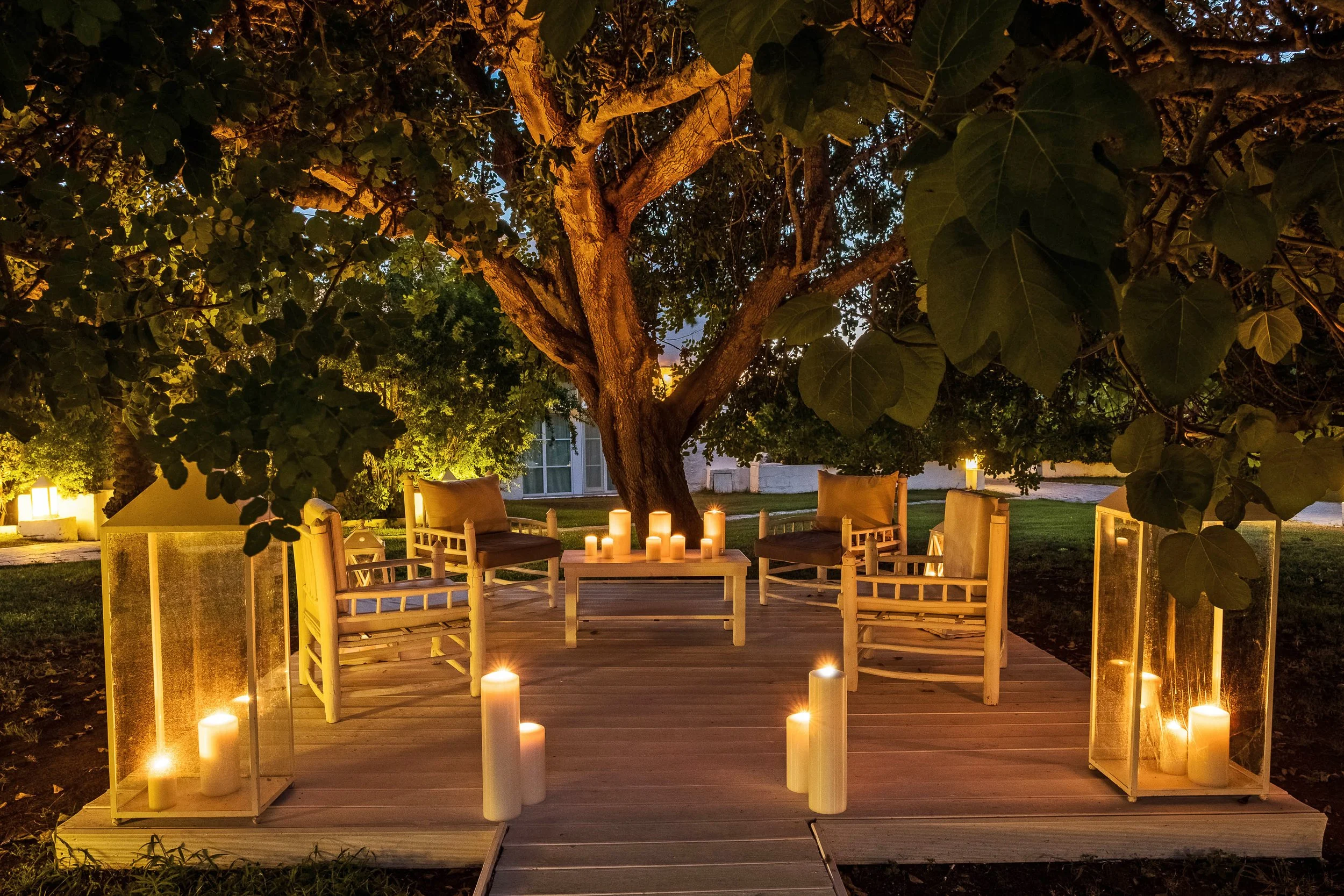Outdoor patio area with seating under a large tree illuminated at night, candles scattered on the deck and surrounding the space.