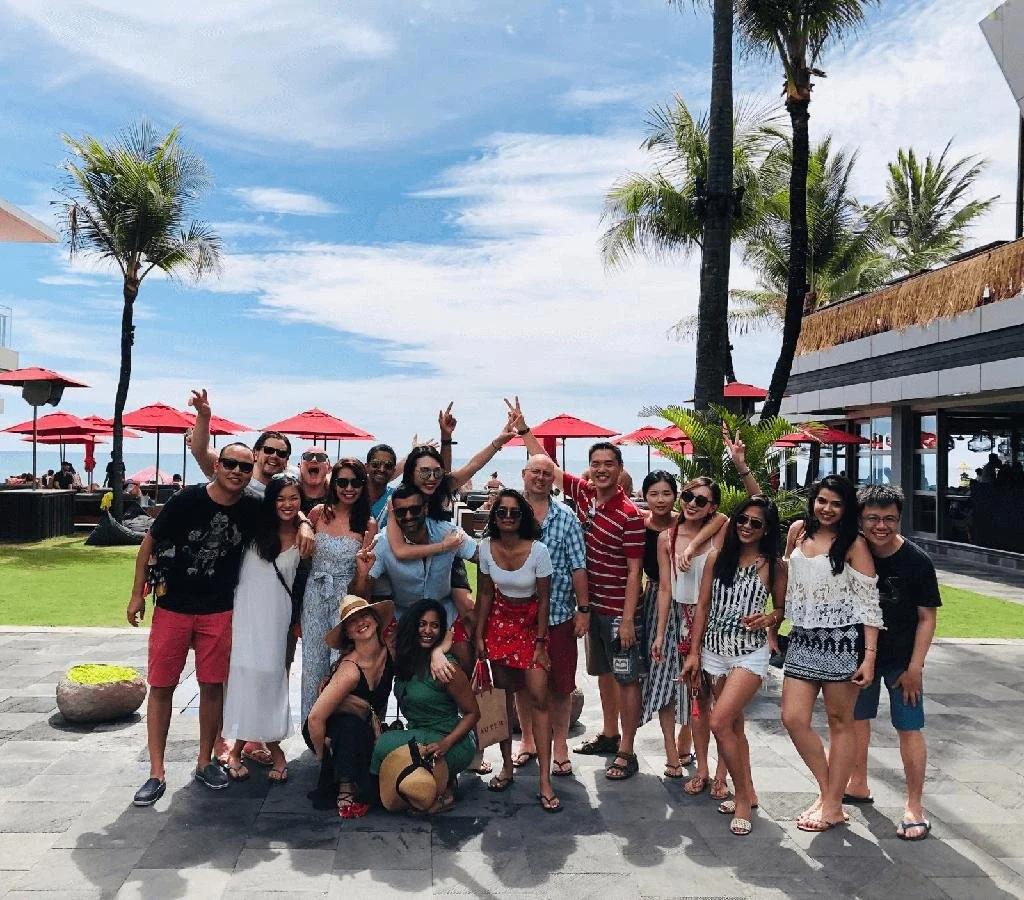 A group of people posing outdoors on a sunny day at a beachside resort, with palm trees, red umbrellas, and a building in the background.