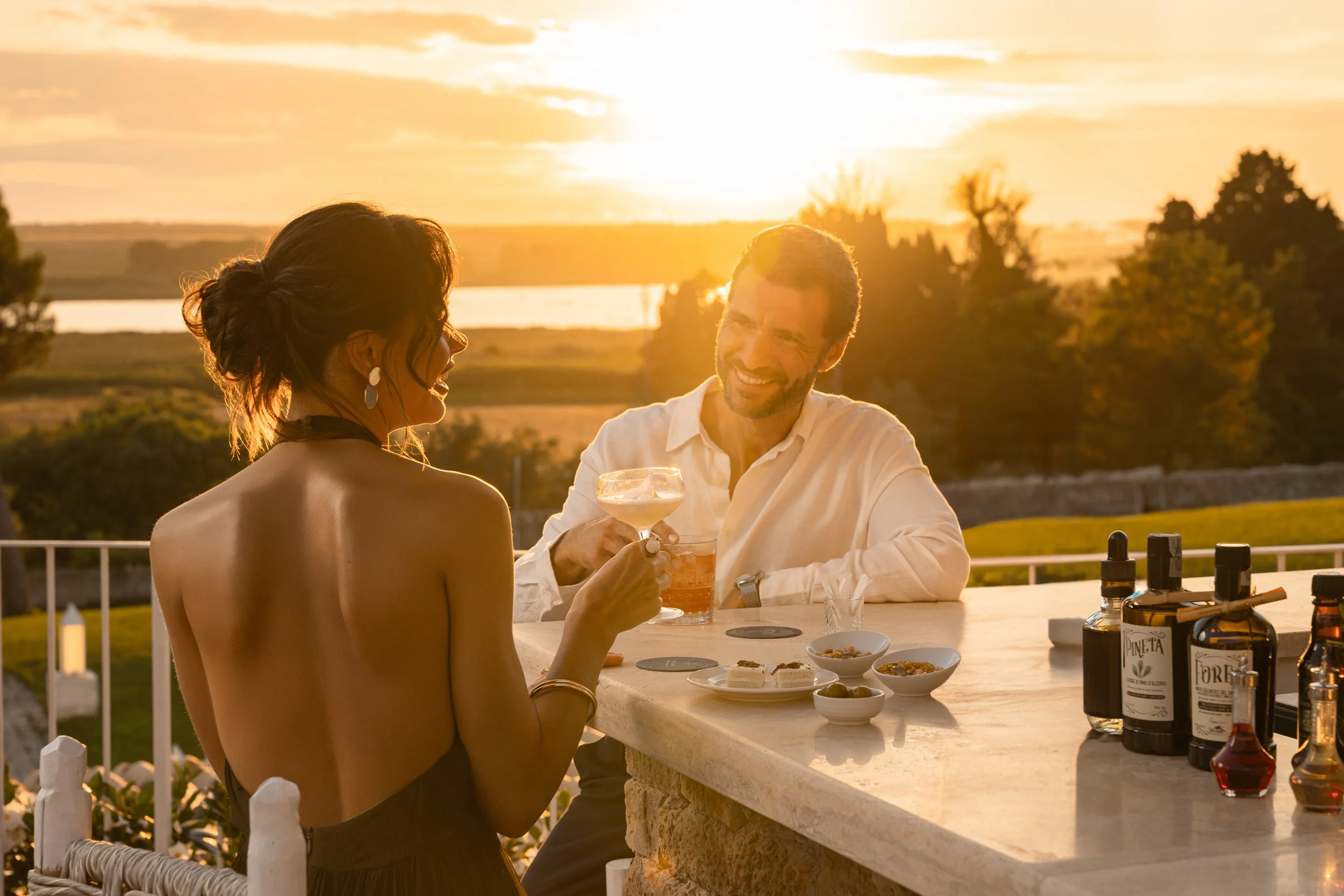 A woman and man enjoying drinks at an outdoor table during sunset. The woman has dark hair in an updo, wears earrings, and a strapless dress. The man has short hair and a beard, wearing a white shirt. The sky is golden with a bright sun, and there are trees and a river in the background. On the table are food bowls, bottles, and glasses.