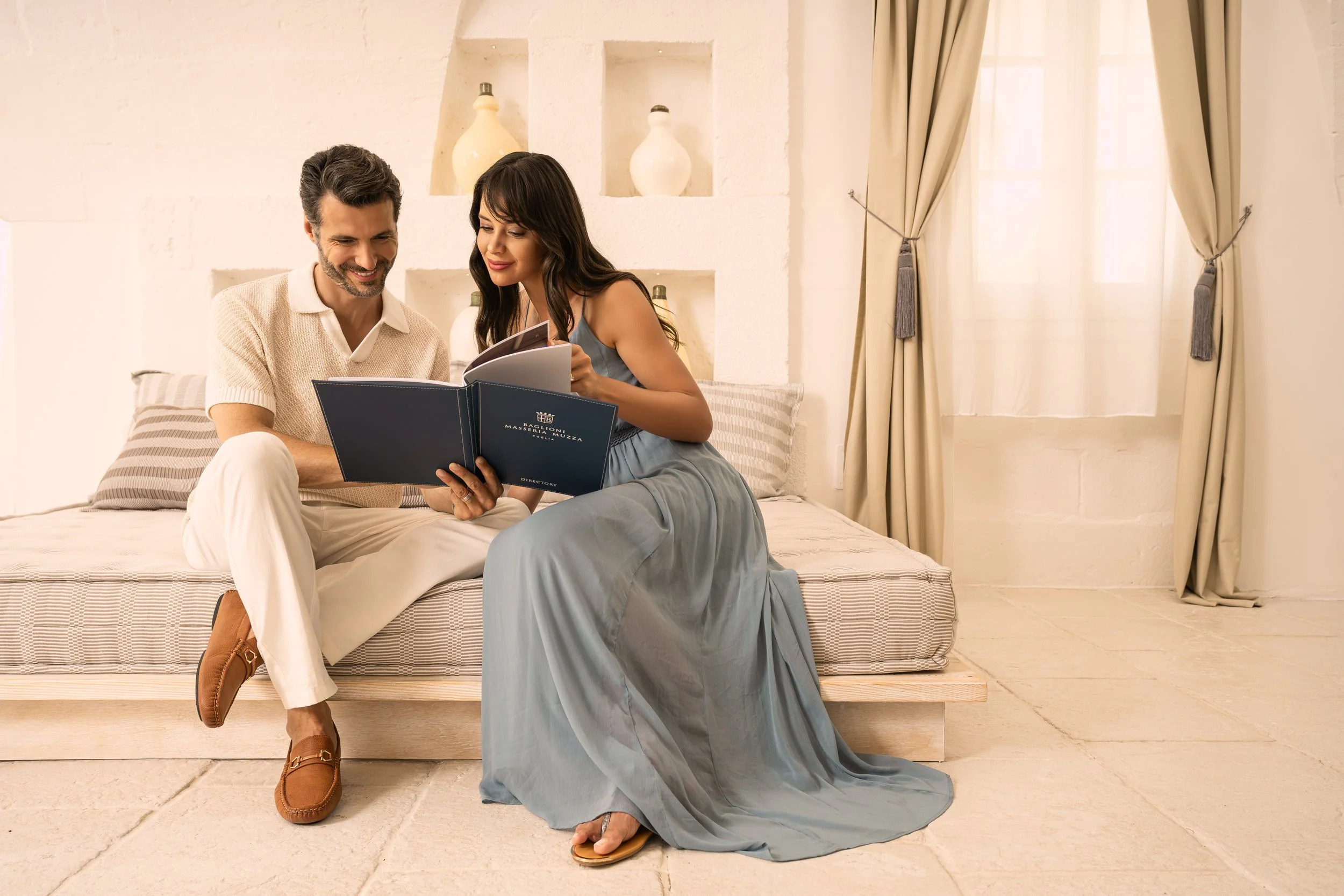 A man and woman sitting on a bed, looking at a photo album together, in a bright, softly decorated room with curtains and decorative vases.