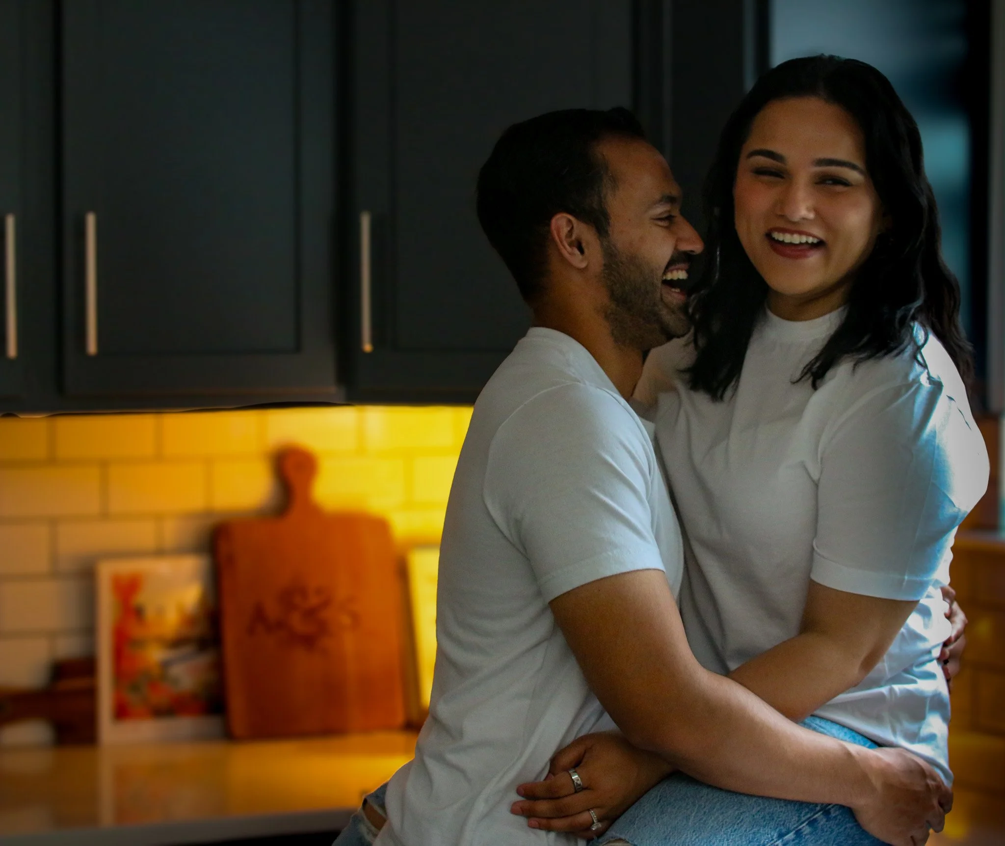 Two people smiling affectionate embrace in a kitchen with dark cabinets and colorful artwork in the background.