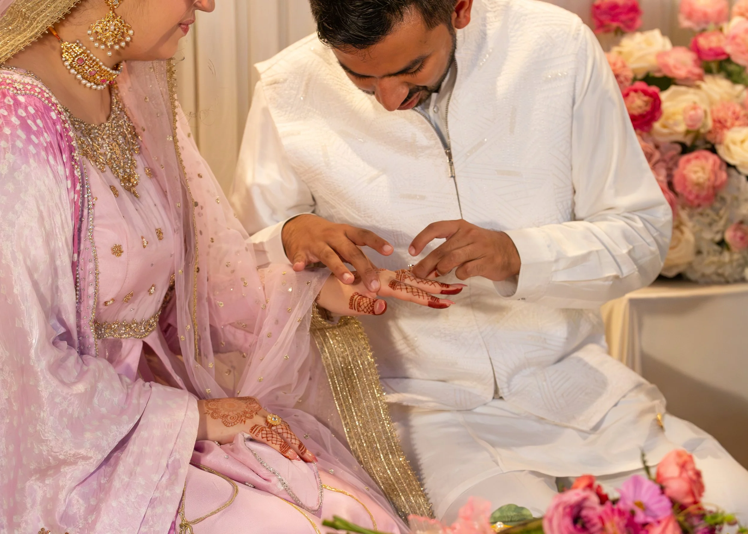A couple traditional Indian wedding ceremony with the groom placing a ring on the bride's finger, both dressed in elaborate wedding attire, surrounded by pink and white flowers.