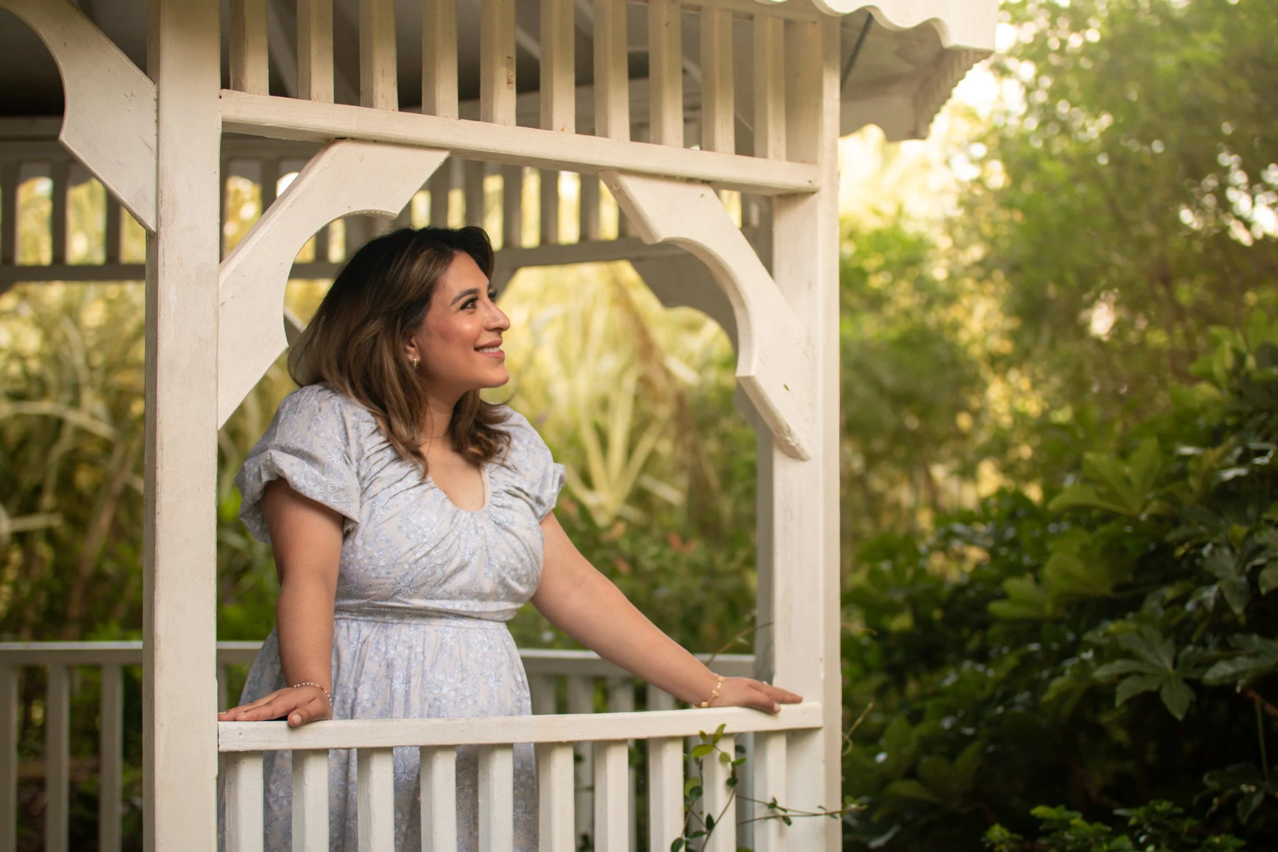 A woman standing on a porch with a white wooden railing, looking to her left and smiling. She is wearing a light patterned dress and is surrounded by greenery.