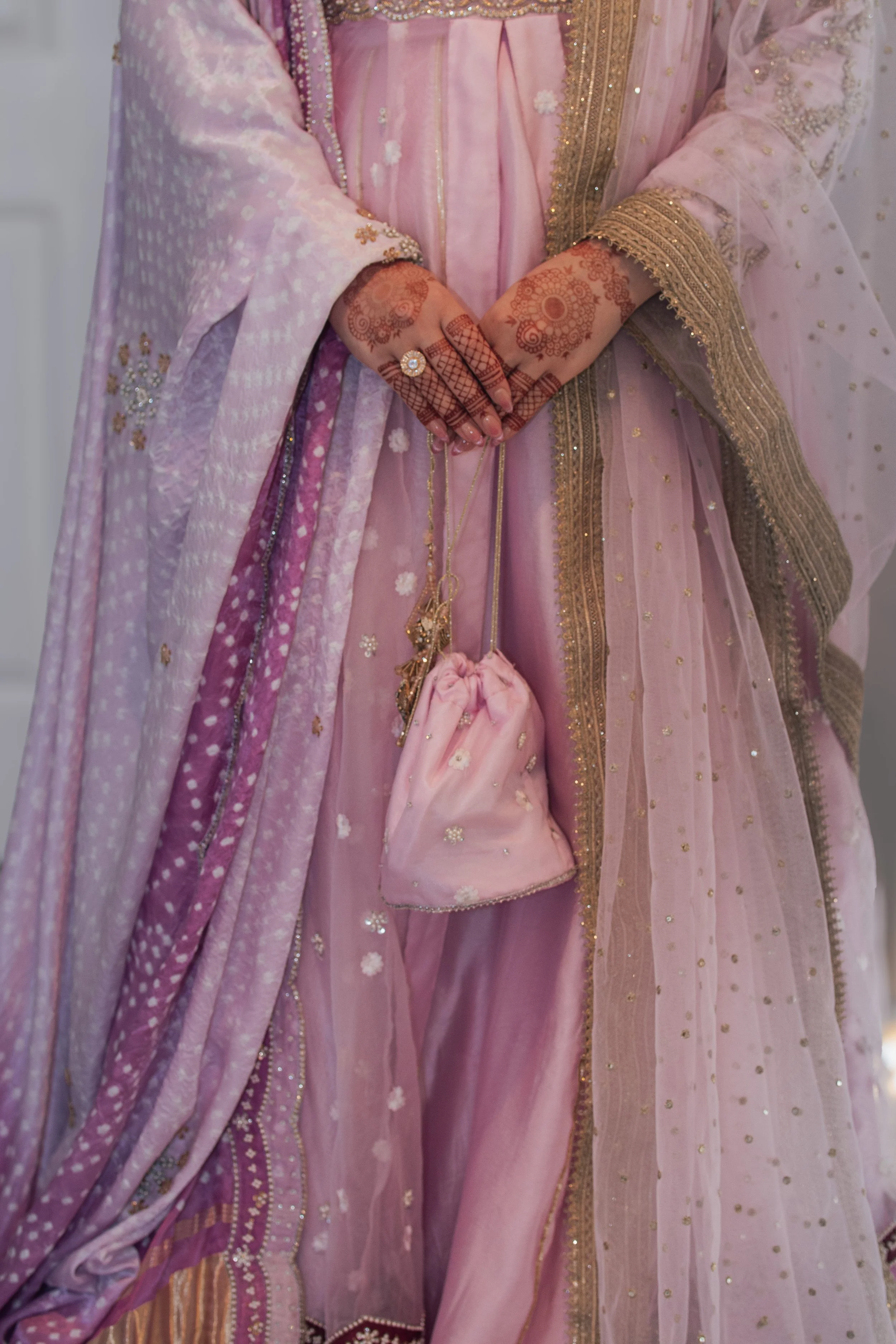 Close-up of a woman wearing a pink traditional saree with gold embroidery. Her hands are decorated with henna designs, and she is holding a matching pink pouch.