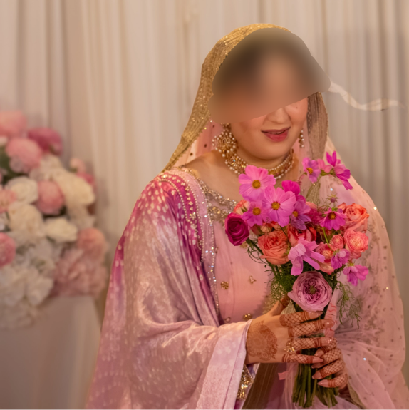 A woman dressed in pink traditional attire with jewelry, holding a bouquet of pink and purple flowers, standing near a floral arrangement.