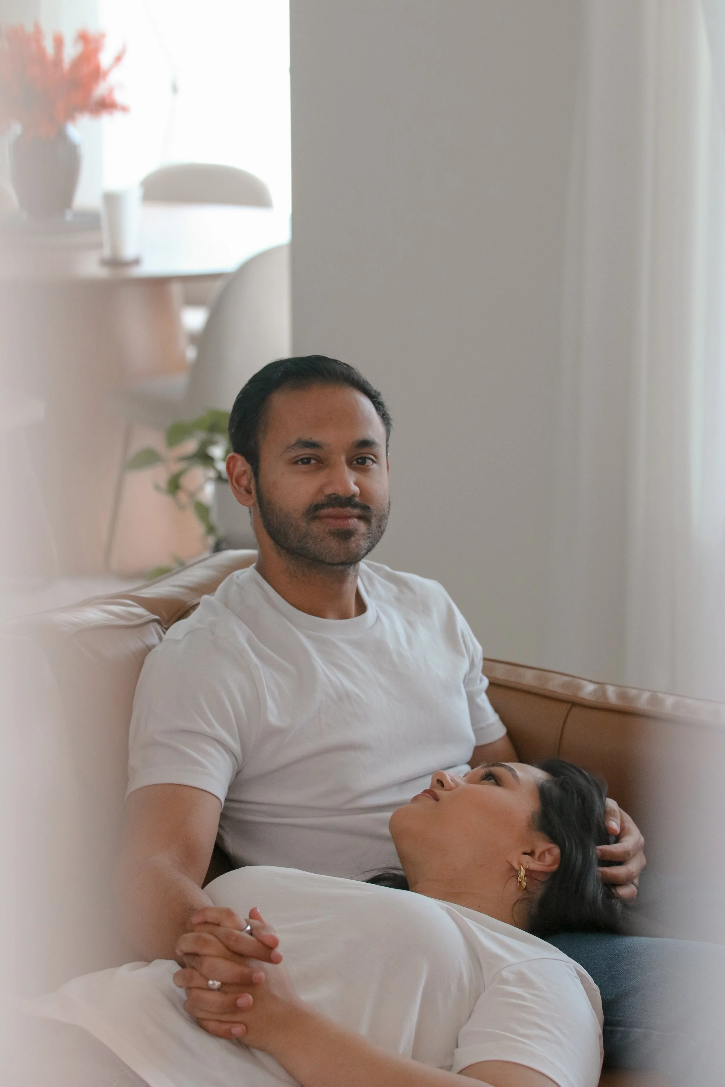 A man with dark hair and beard sitting on a beige couch, holding a woman lying on her back. The woman has dark hair, is wearing earrings, and is gazing up at the man. The setting appears to be a living room with a wooden side table, a potted plant, and a vase with flowers in the background, with natural light coming through a window.