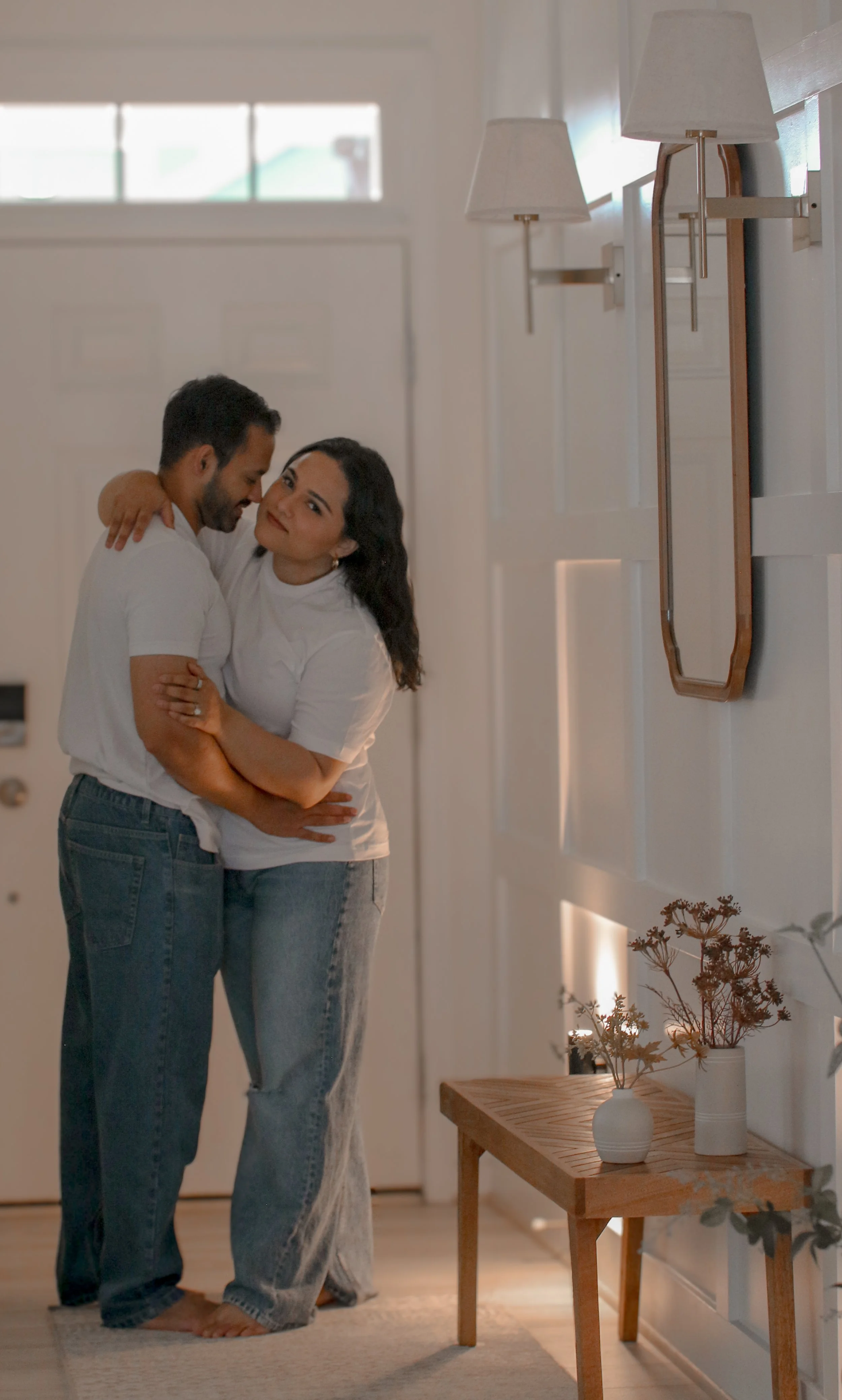 A man and woman hugging in a cozy, well-lit living room with a wall mirror, wall-mounted lamps, and a small wooden table with decorative vases and dried flowers.