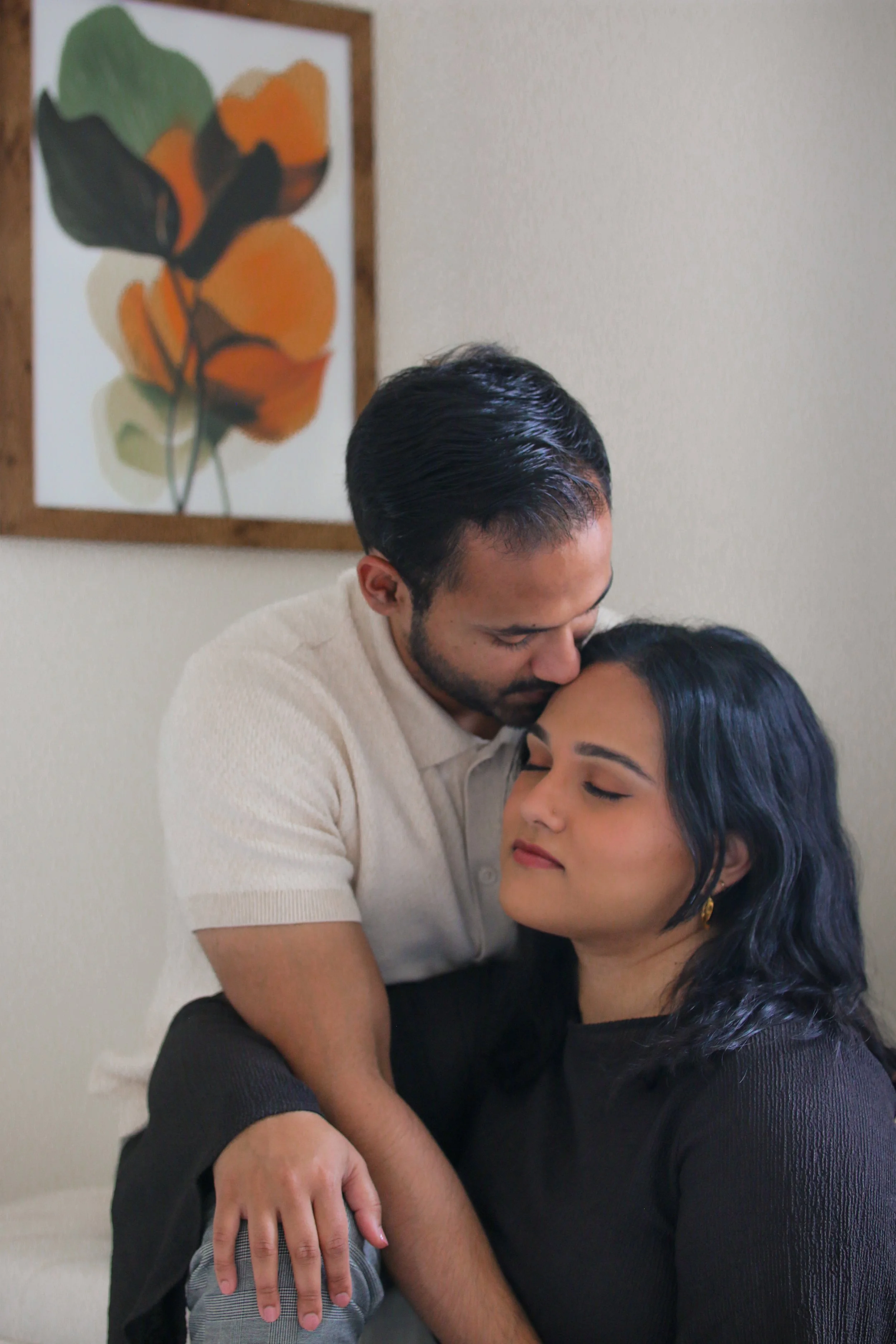 A man is hugging and leaning his head on a woman with long dark hair, as she closes her eyes and smiles softly. The scene is indoors with a neutral-colored wall and a framed abstract painting of orange and green shapes in the background.