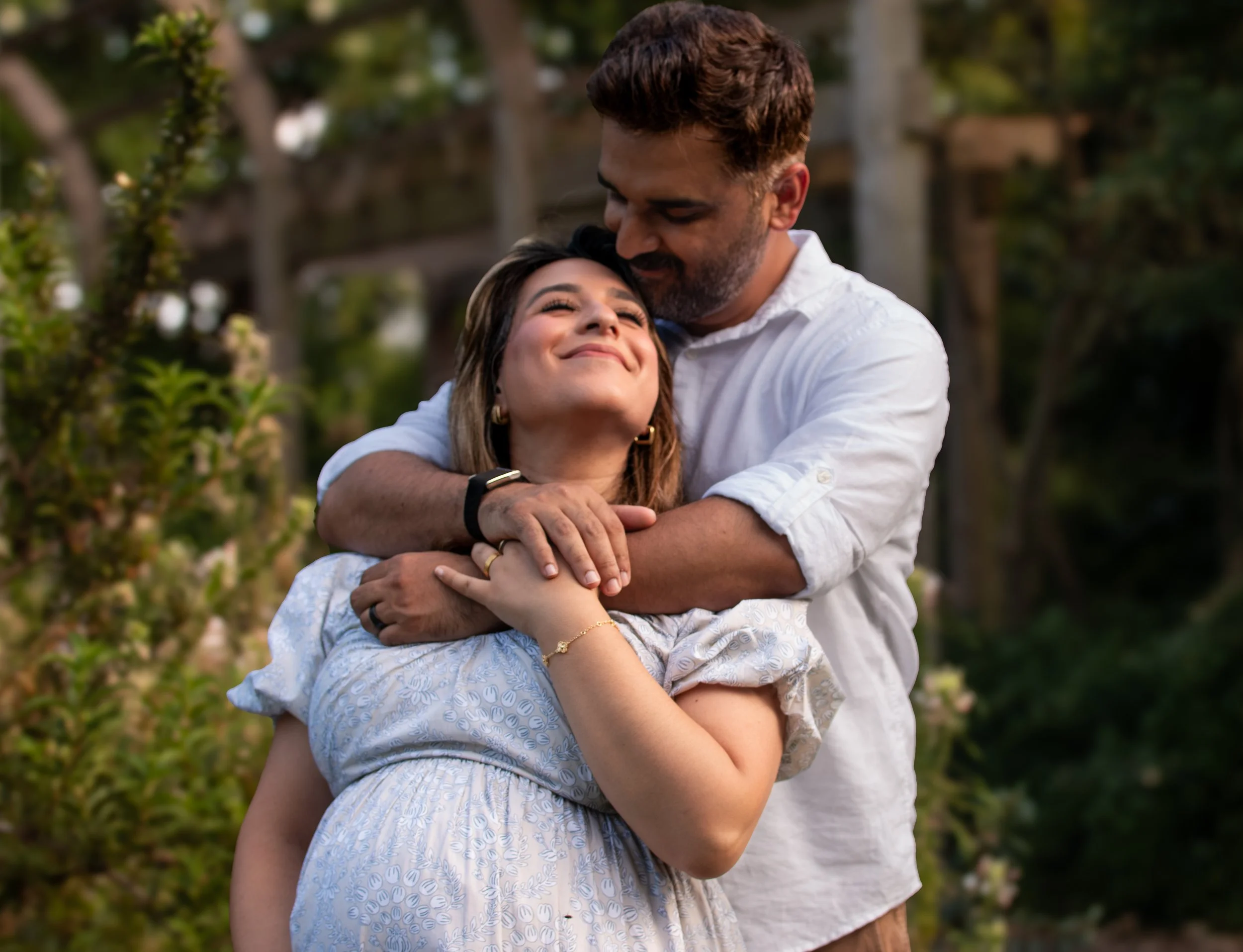 A man is hugging a woman in an outdoor garden setting. The woman has long hair and is smiling with her eyes closed, while the man has short hair and a beard, and is resting his forehead on her head.
