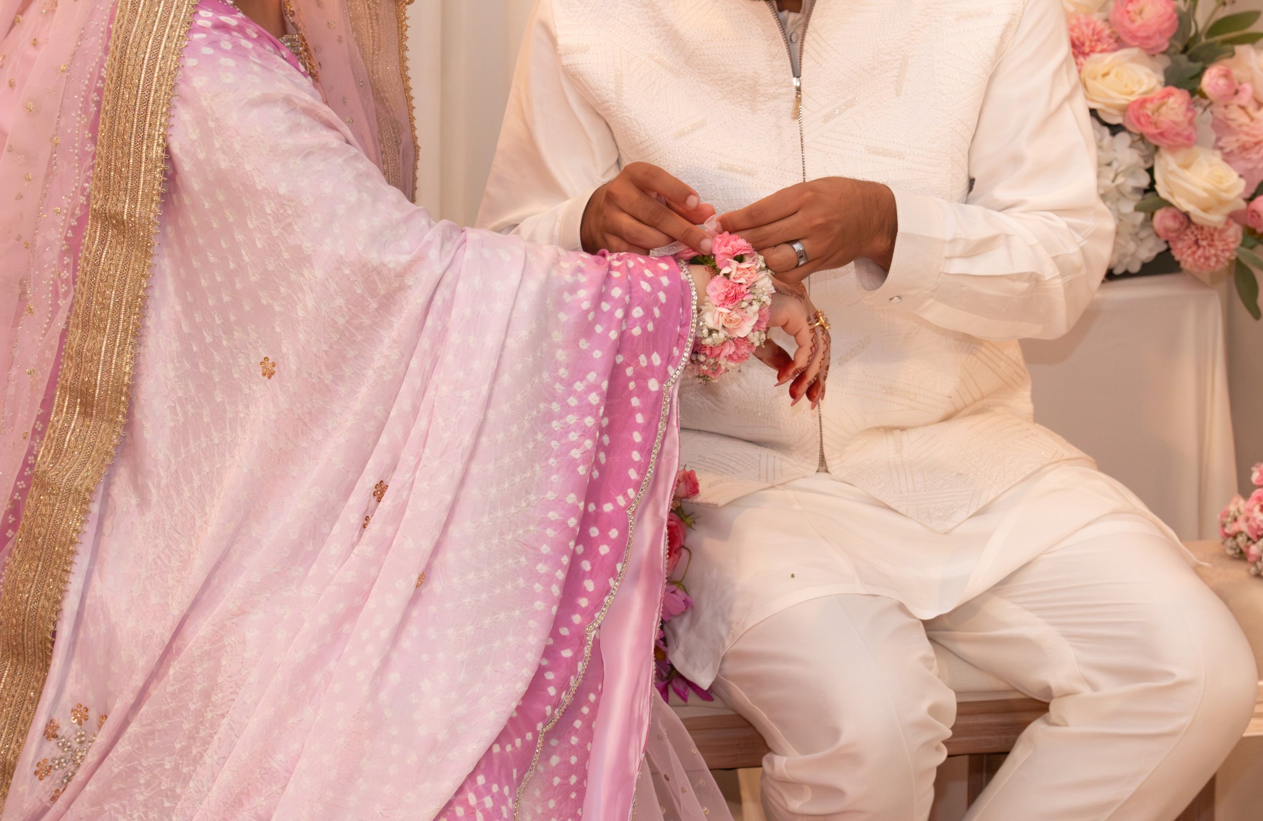 A couple during a wedding ceremony exchanging rings. The woman is wearing a pink sari with gold embroidery, and the man is wearing a cream-colored traditional outfit. Pink and white flowers are present in the background.