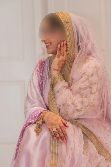 A woman dressed in traditional South Asian attire with intricate henna designs on her hands, smiling and touching her cheek gently.