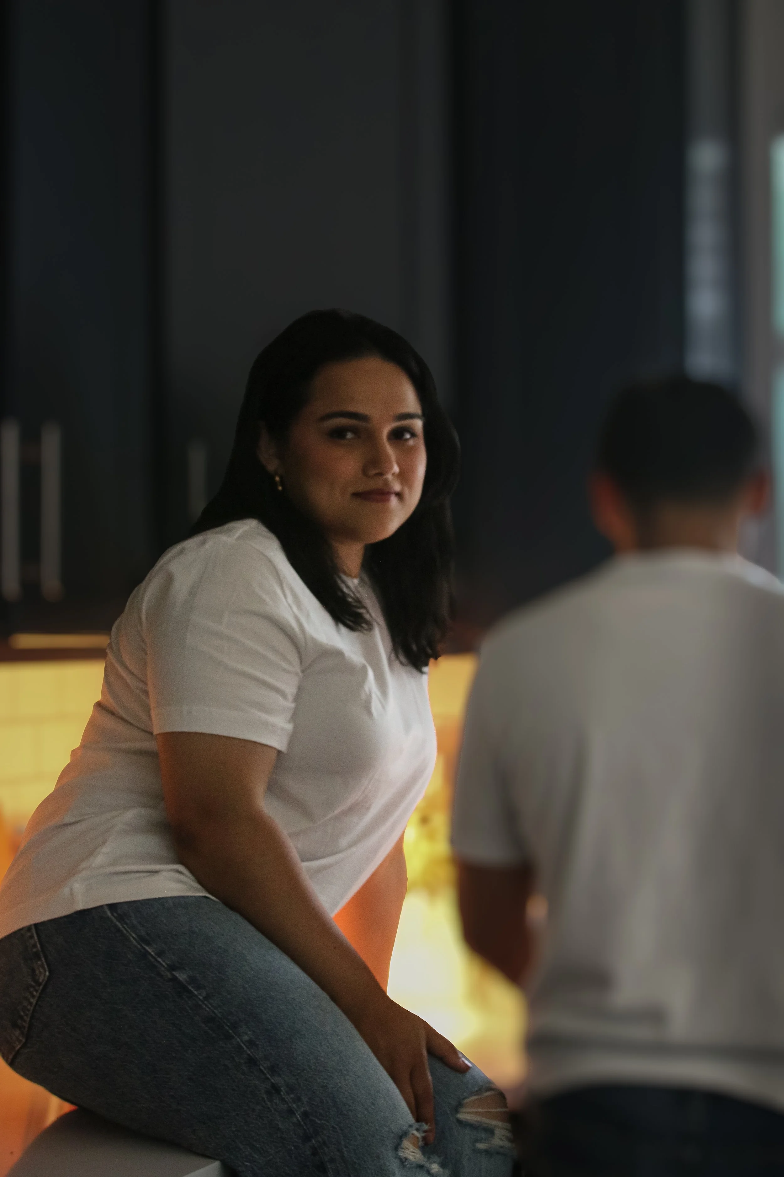 A woman sitting in a conversation, wearing a white shirt and jeans, in a dimly lit indoor setting with dark background and warm lighting.