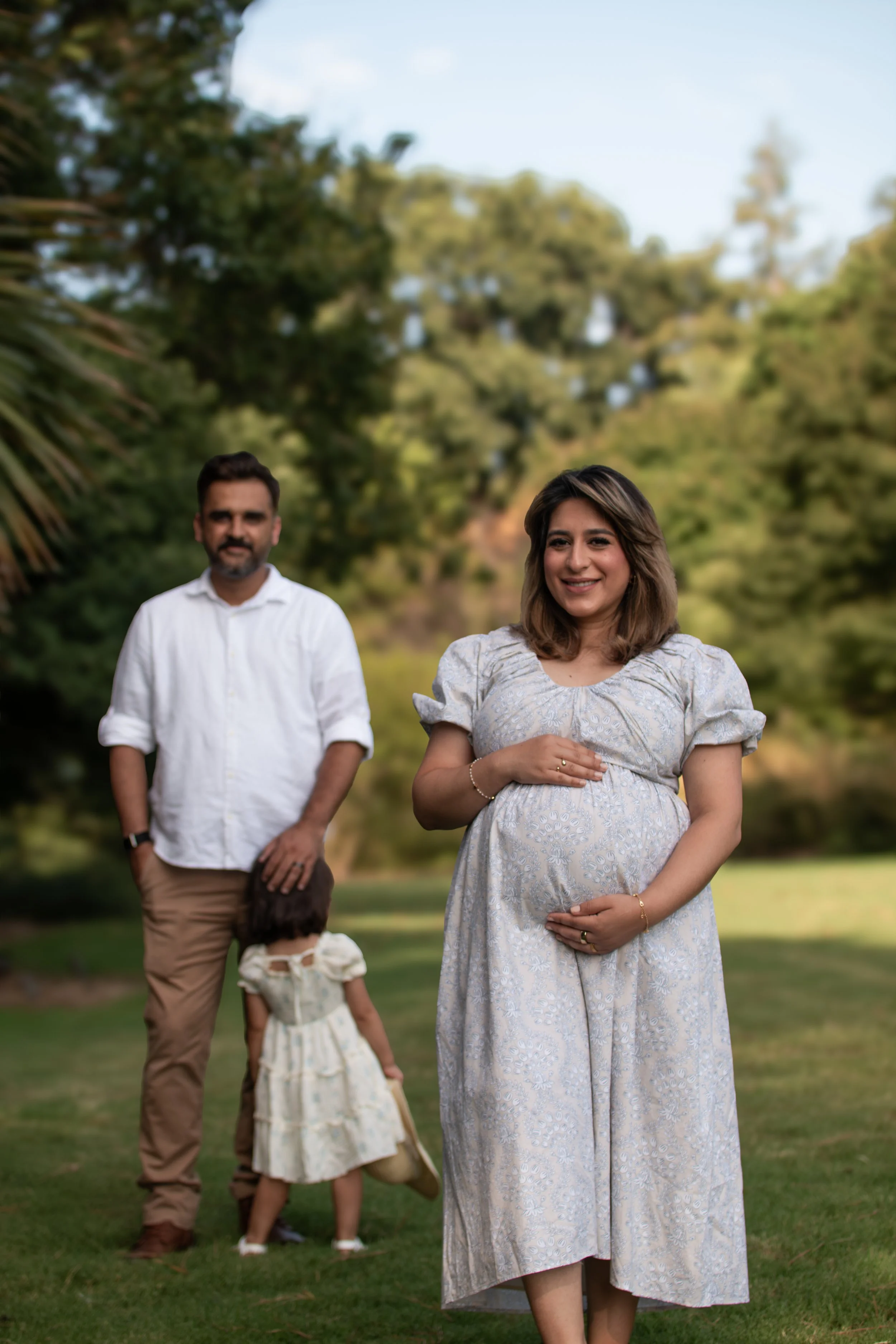 A pregnant woman in a light-colored dress standing outdoors, smiling, with a man and a girl in the background on a grassy area with trees.