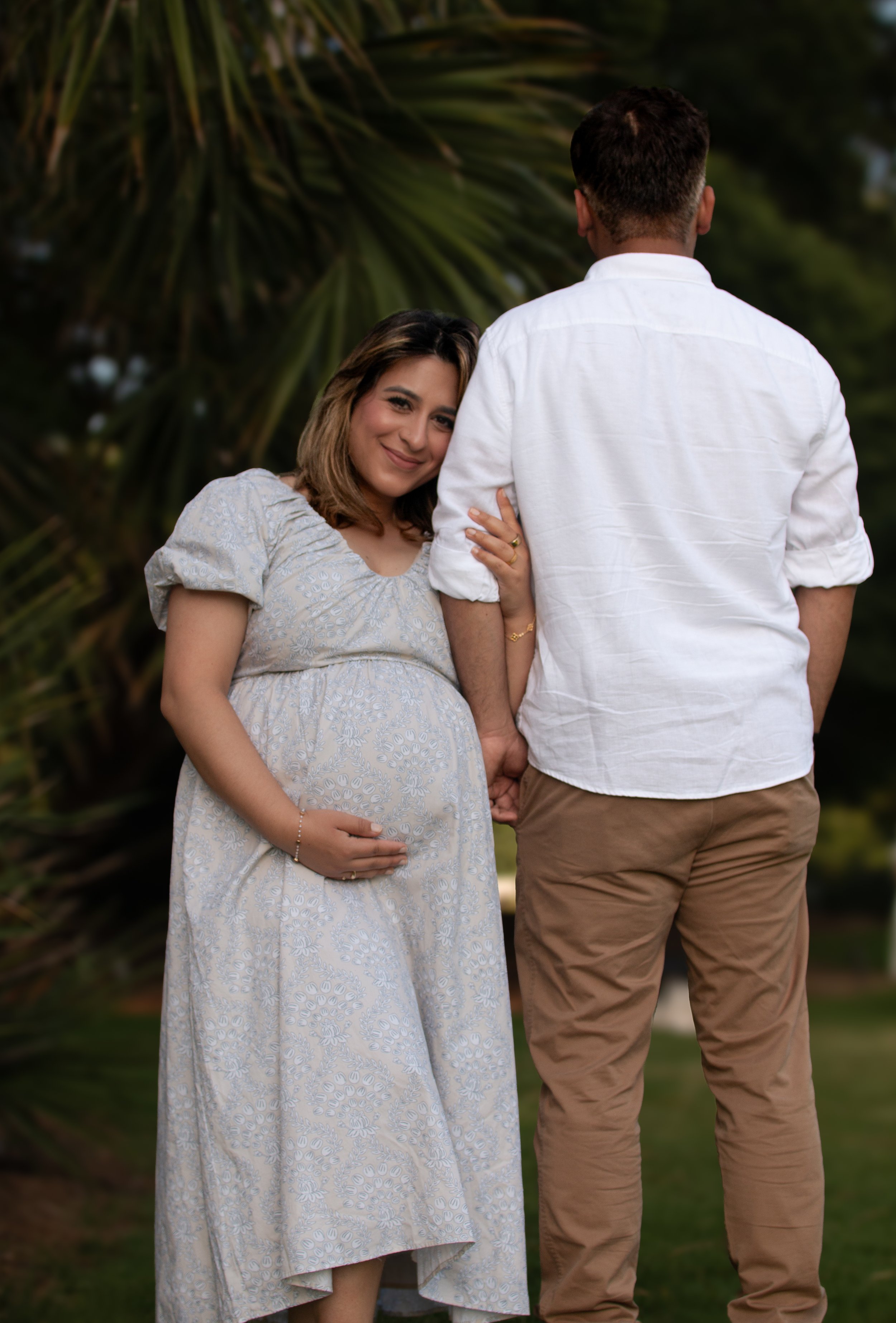 A pregnant woman smiling and resting her head on a man’s shoulder outdoors. The woman wears a light-colored dress with floral patterns, and the man wears a white shirt and beige pants. They are holding hands, with a background of green foliage.