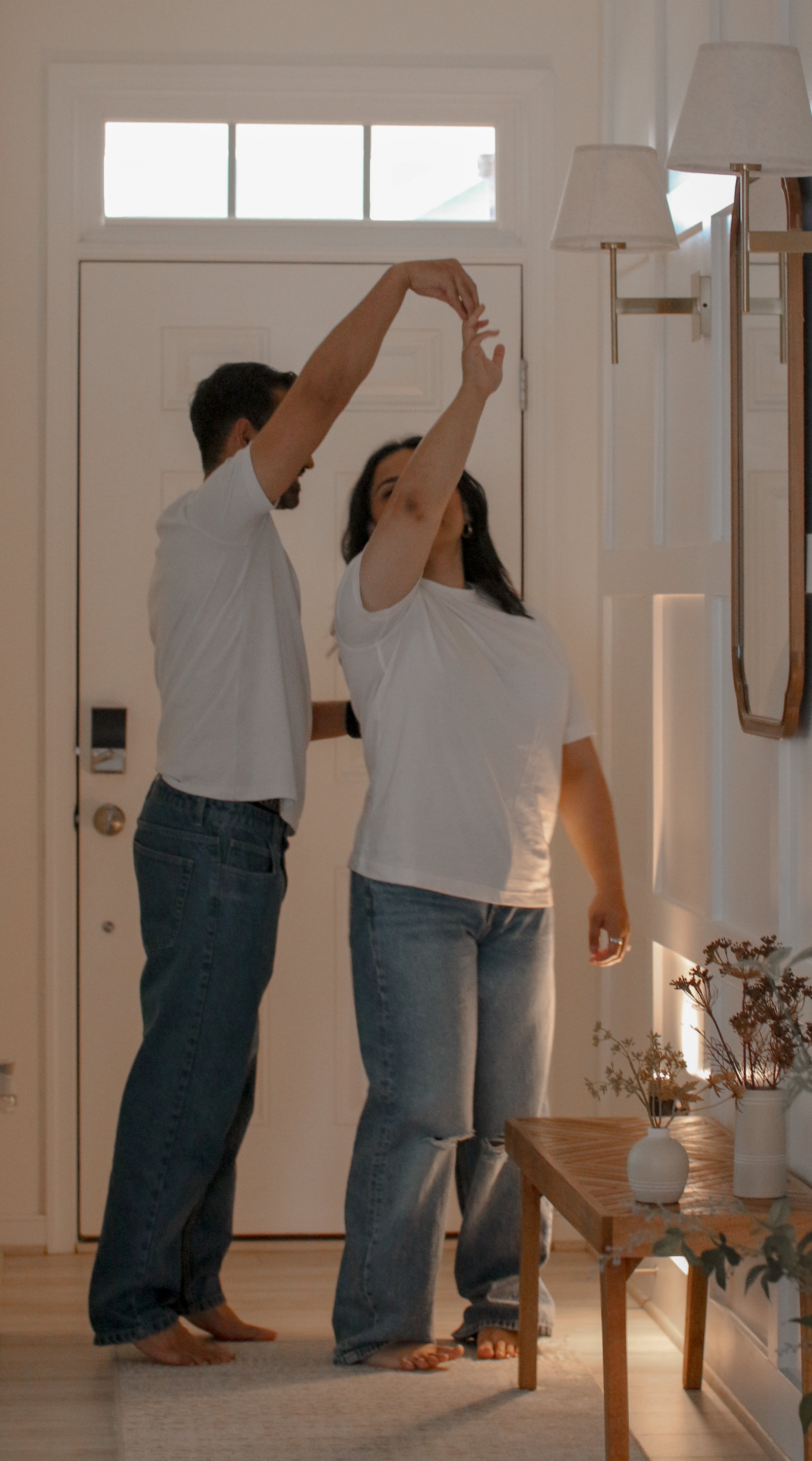 Two people, a man and a woman, standing inside a house near the front door, with the man holding the woman's hand up in a dance pose. Both are dressed casually in white T-shirts and jeans, and the scene features warm, soft lighting from a nearby wall sconce.