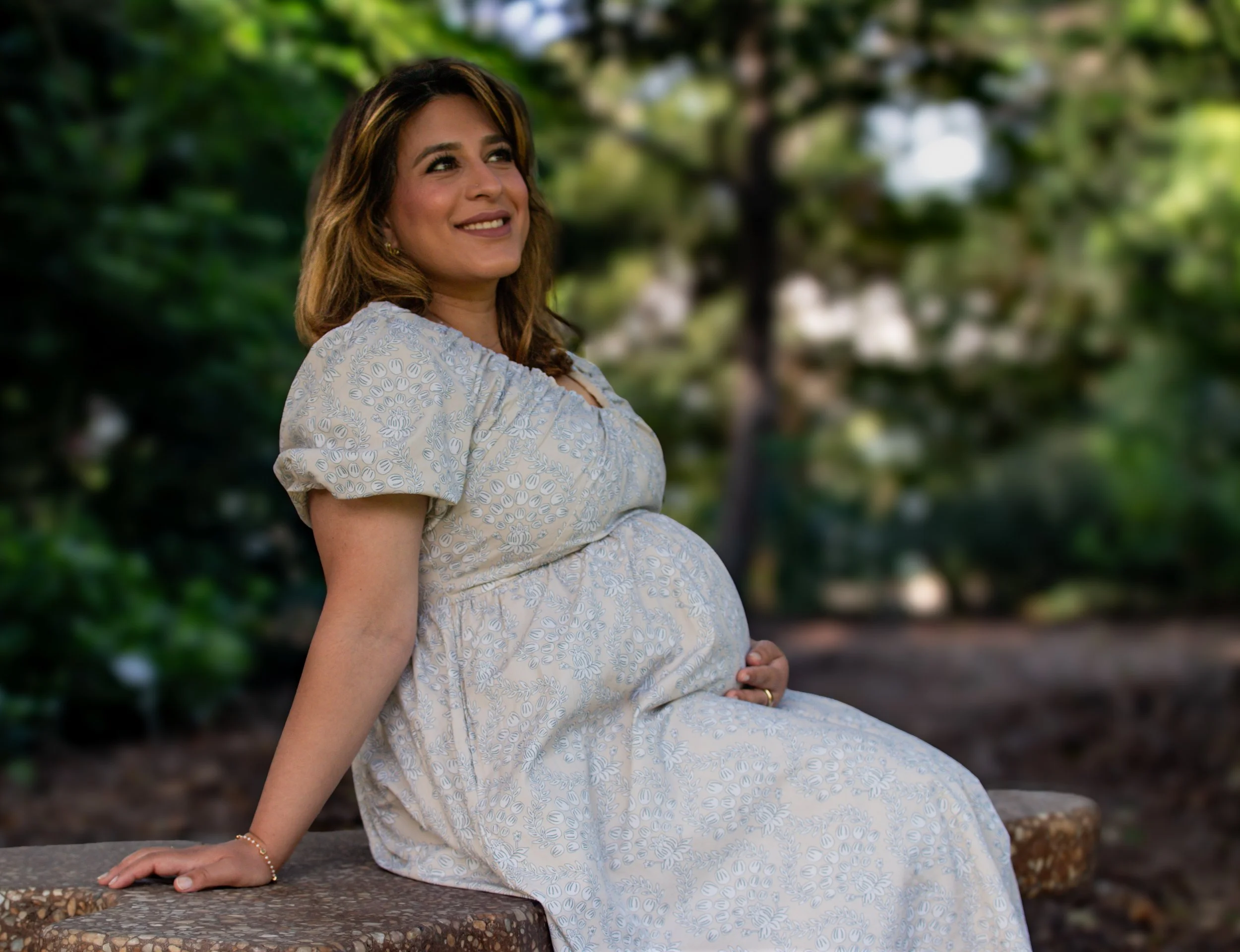 A pregnant woman with light brown hair and a warm smile, sitting outdoors on a bench, wearing a light-colored floral dress, with greenery in the background.