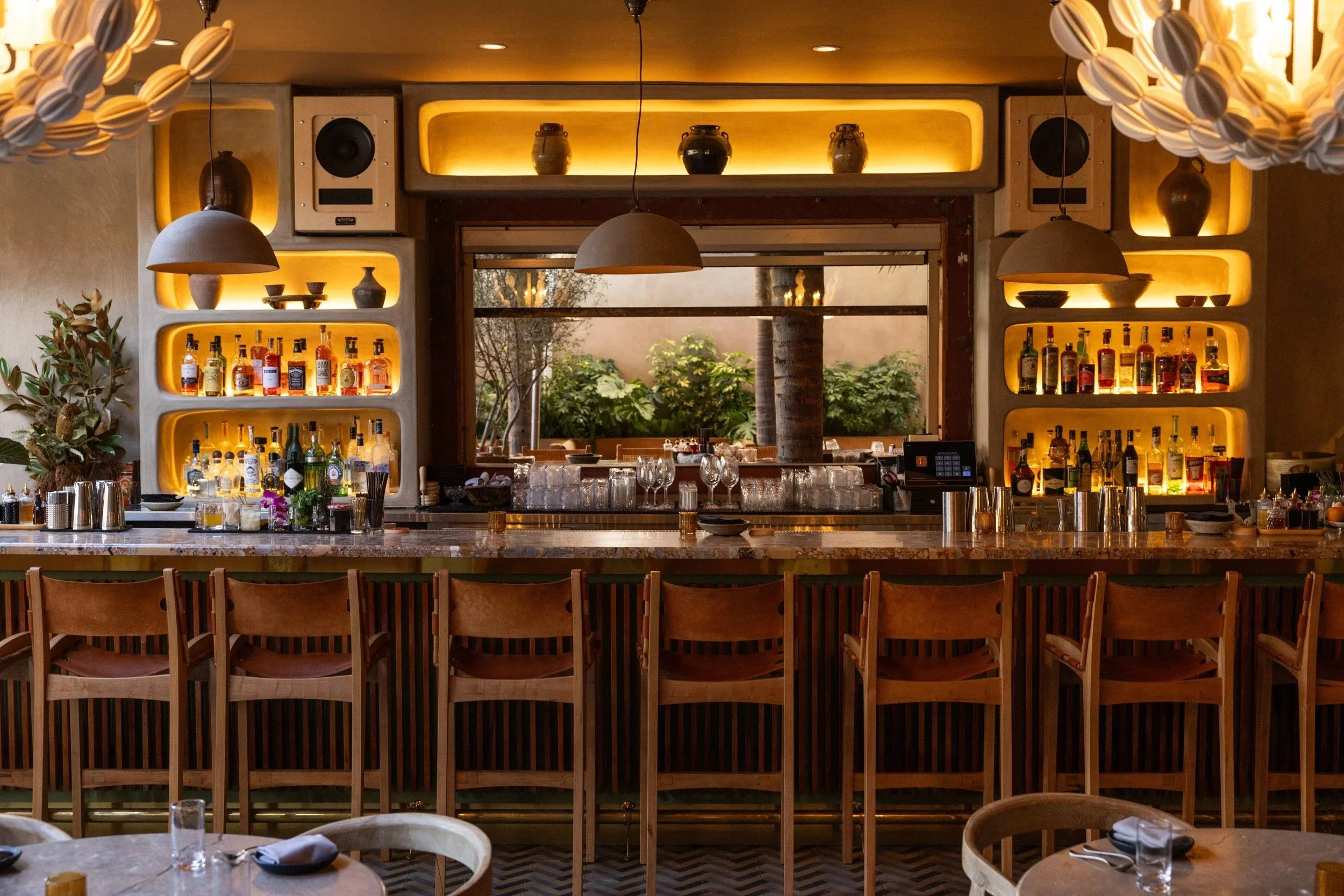 Interior view of a restaurant bar area with a marble countertop, wooden chairs, and shelves displaying bottles of liquor. Decor includes hanging pendant lights and vases on illuminated shelves. Large window showing outdoor greenery.